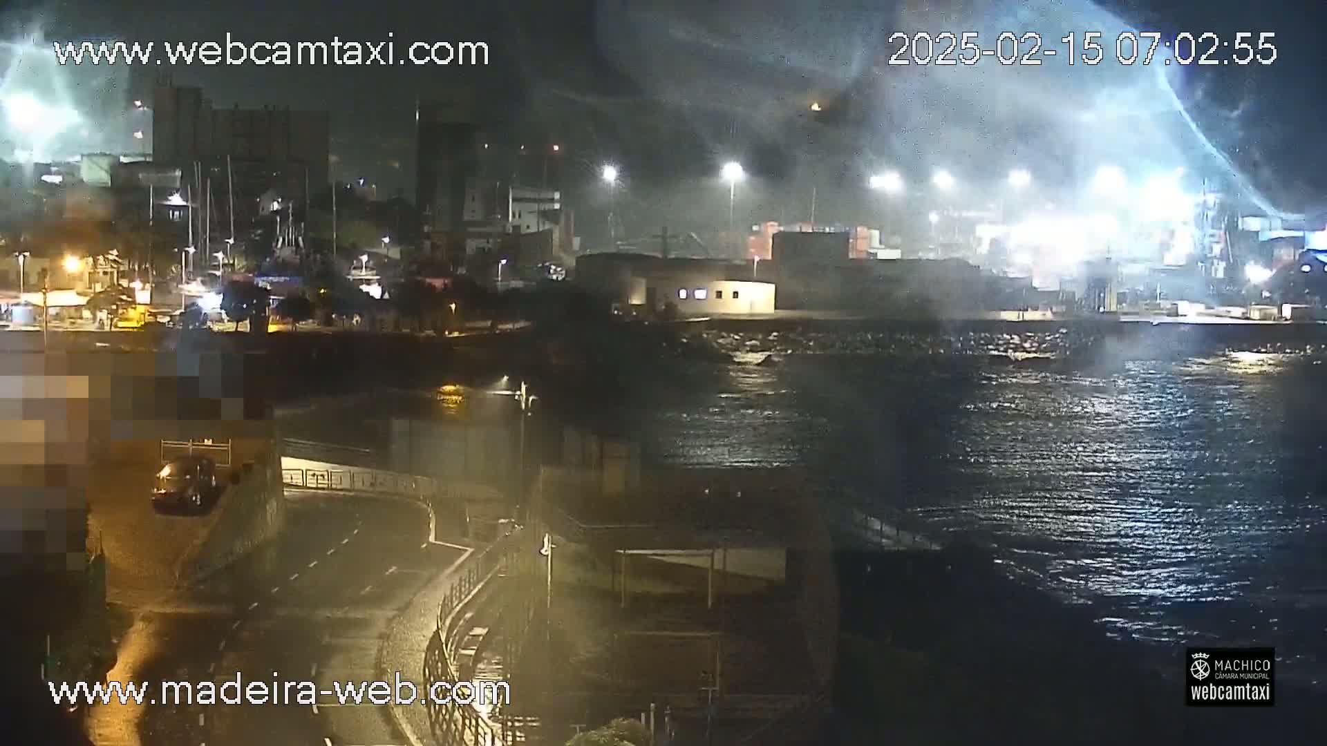 A nighttime view of a harbor with choppy water under a dark, possibly stormy sky, showing lights illuminating a waterfront road and buildings.