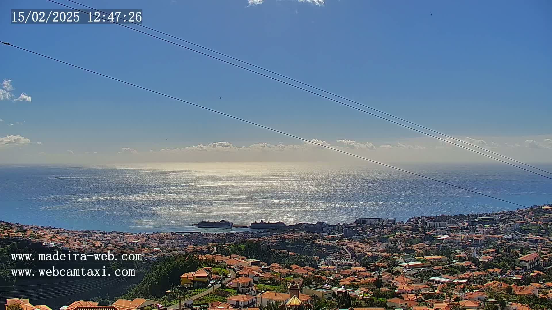 A coastal town with red-tiled roofs sits below a mostly clear blue sky, overlooking a calm ocean reflecting sunlight.