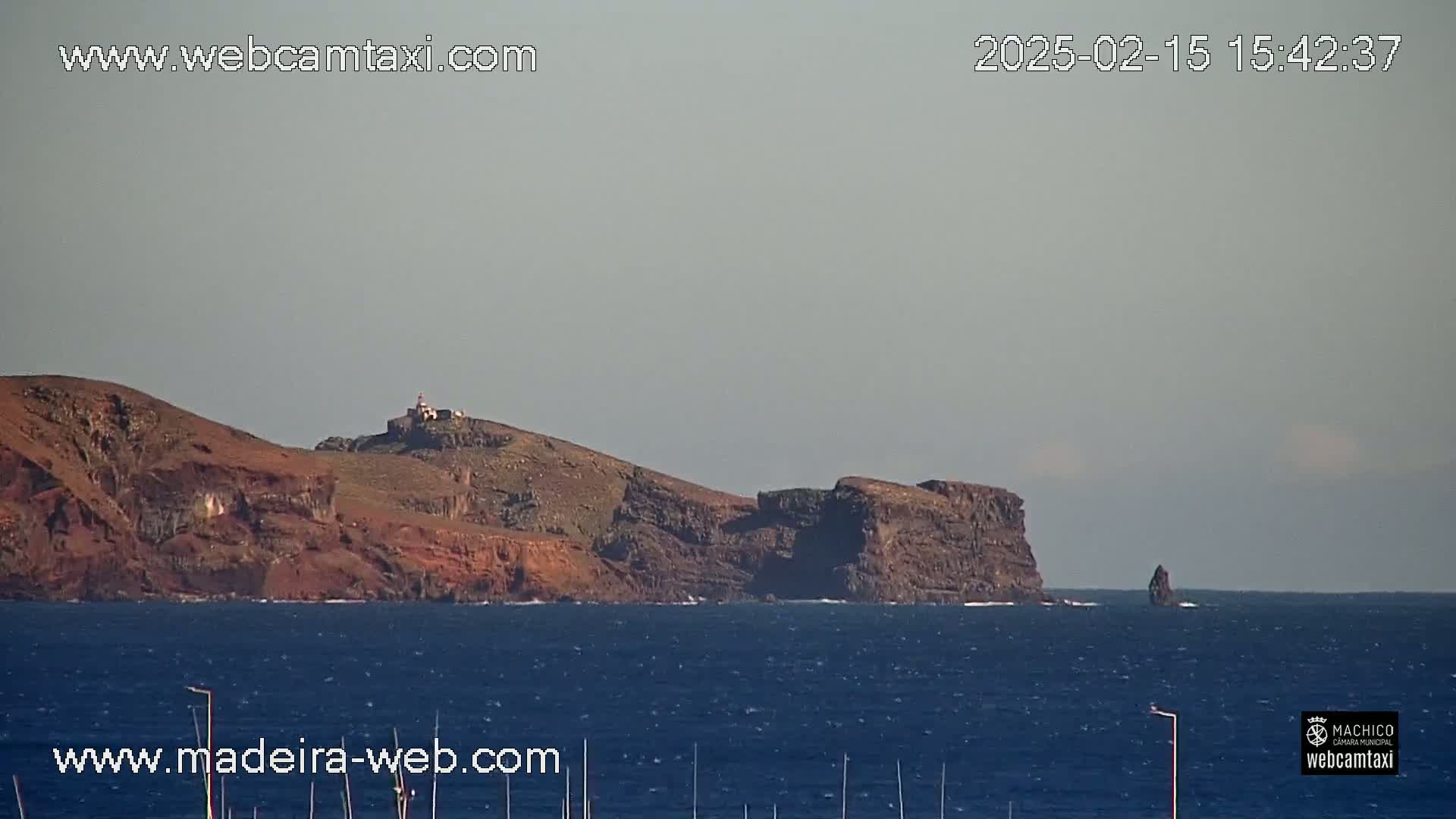 A rocky island with a lighthouse is visible across a calm ocean under a hazy sky.