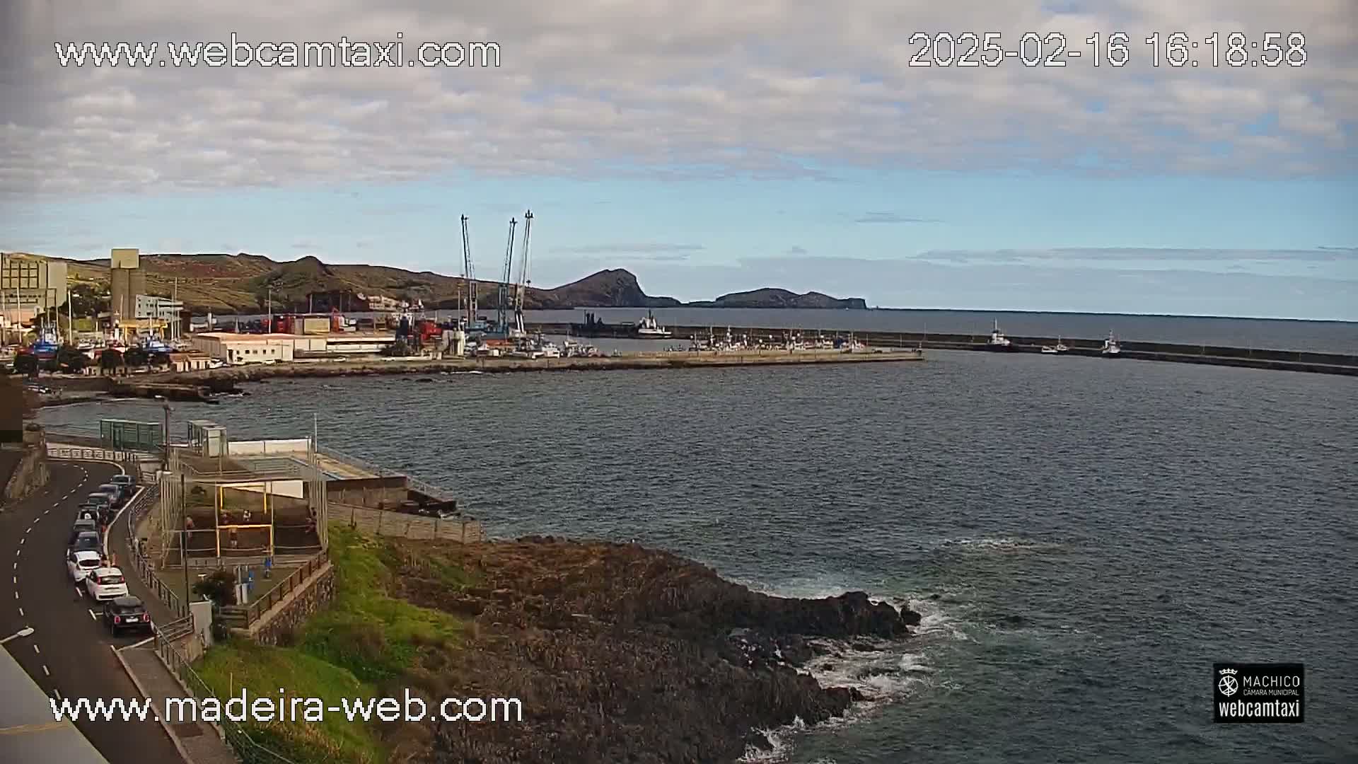 A partly cloudy day reveals a harbor filled with boats, industrial buildings, and a rocky coastline with a calm sea.