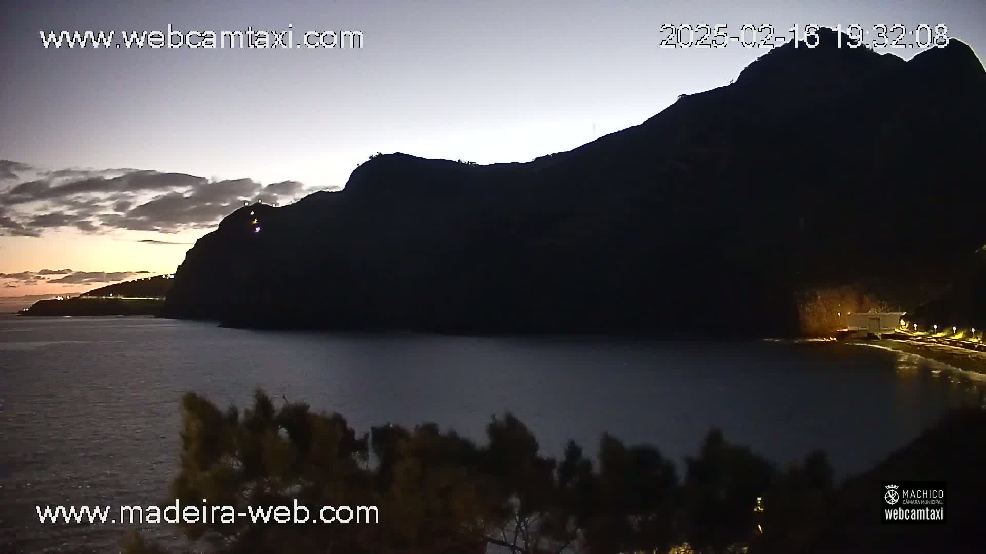 A dark, silhouetted coastal cliff overlooks a calm body of water at twilight under a mostly clear sky.