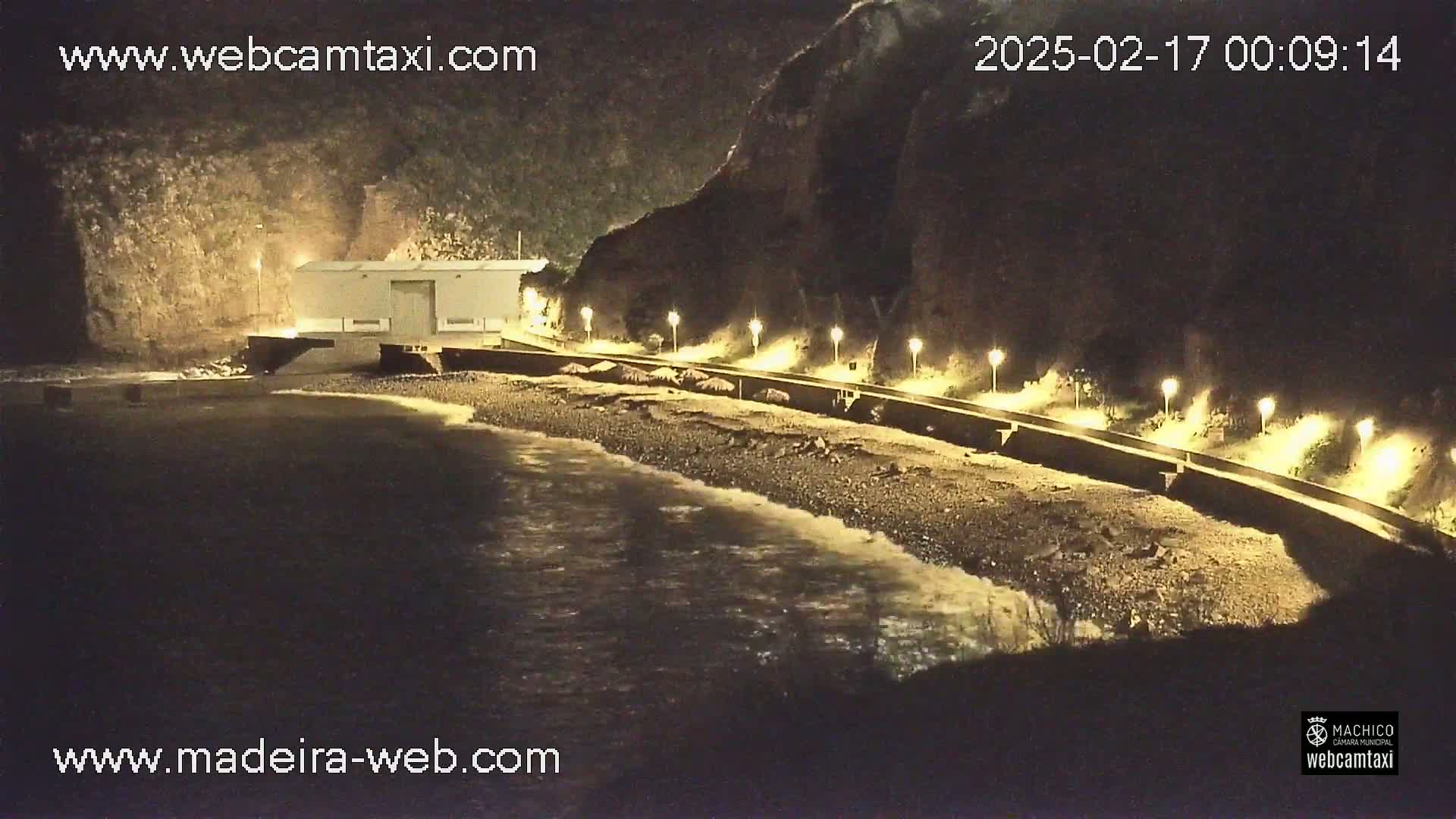 A nighttime view of a dark, rocky beach illuminated by lamps along a seawall, with gentle waves lapping the shore near a small building.