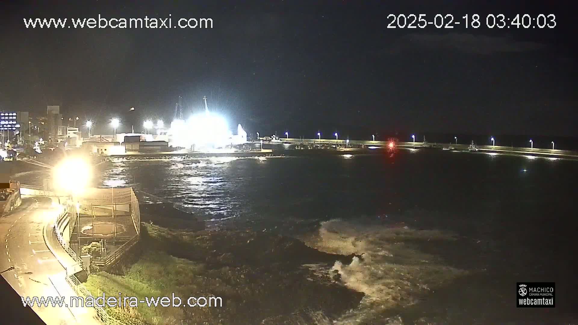 A nighttime view of a harbor with choppy water crashing against a rocky shore, illuminated by lights from buildings and structures along the waterfront.