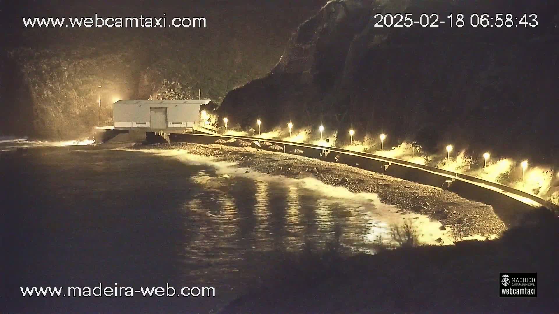 A nighttime view of a rocky coastline with a building near the water, lit by several lamps along a wall, with gentle waves lapping the shore.