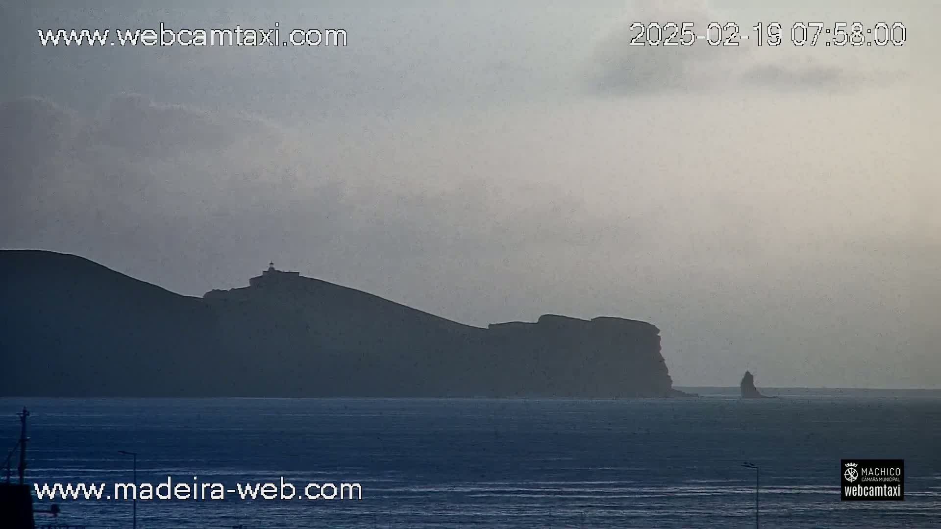 A hazy, overcast day reveals a dark, mountainous coastline with a lighthouse visible on a distant hill, overlooking a calm body of water.