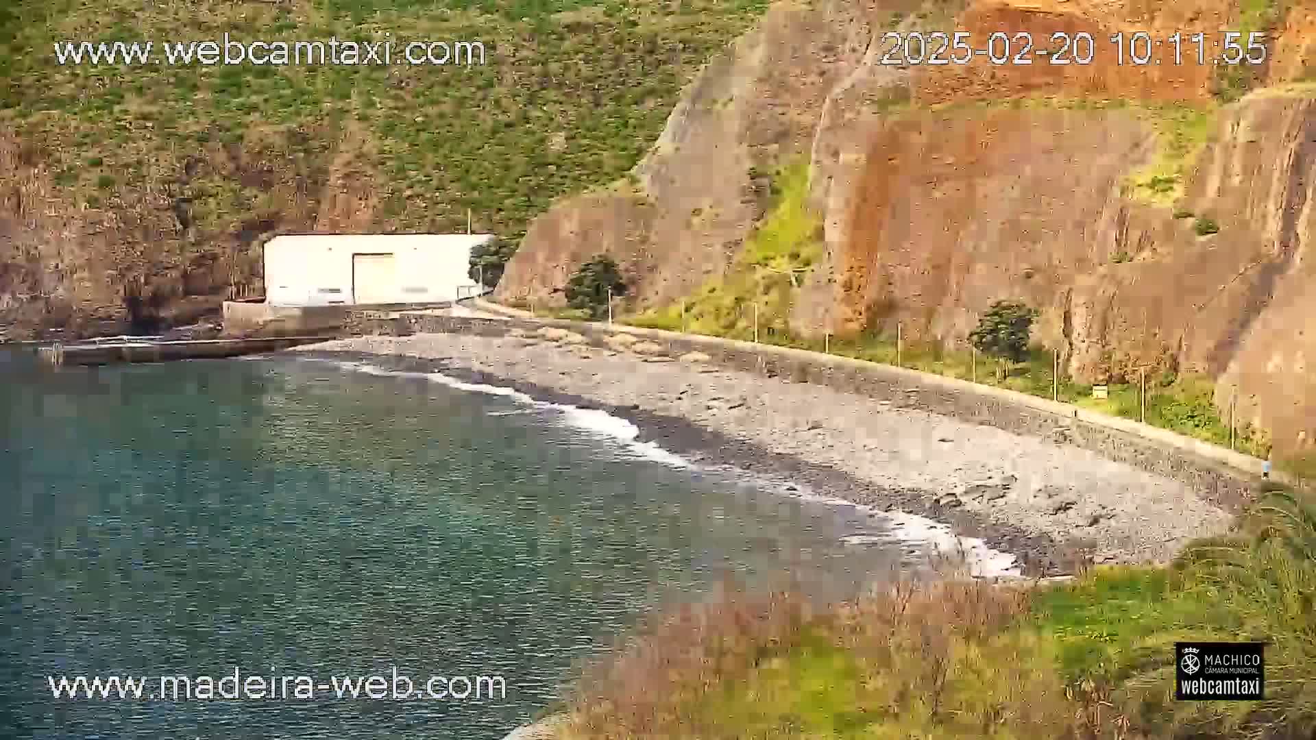 A small, rocky beach with gentle waves laps against the shore next to a white building and a cliffside, under sunny conditions.