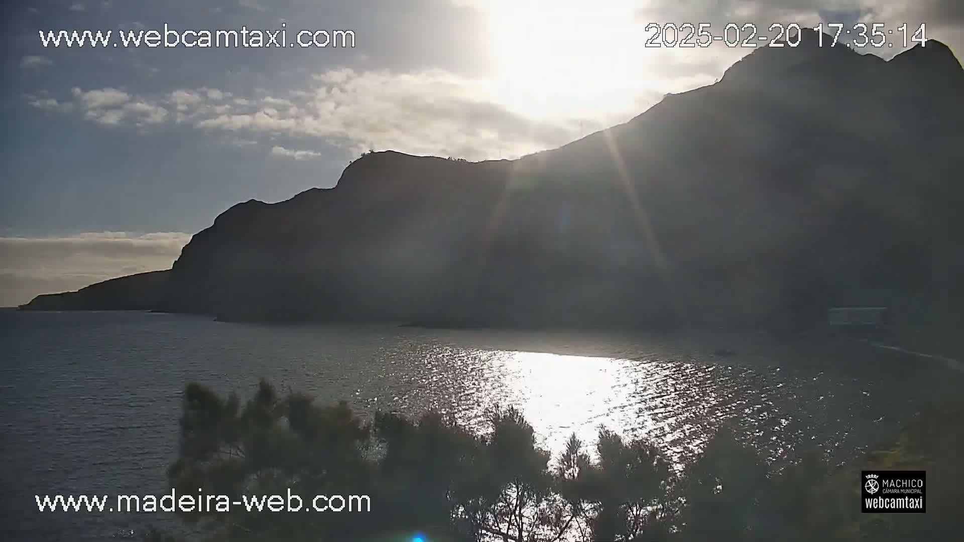 A partly cloudy sunset shines on a calm body of water next to a dark, mountainous shoreline.