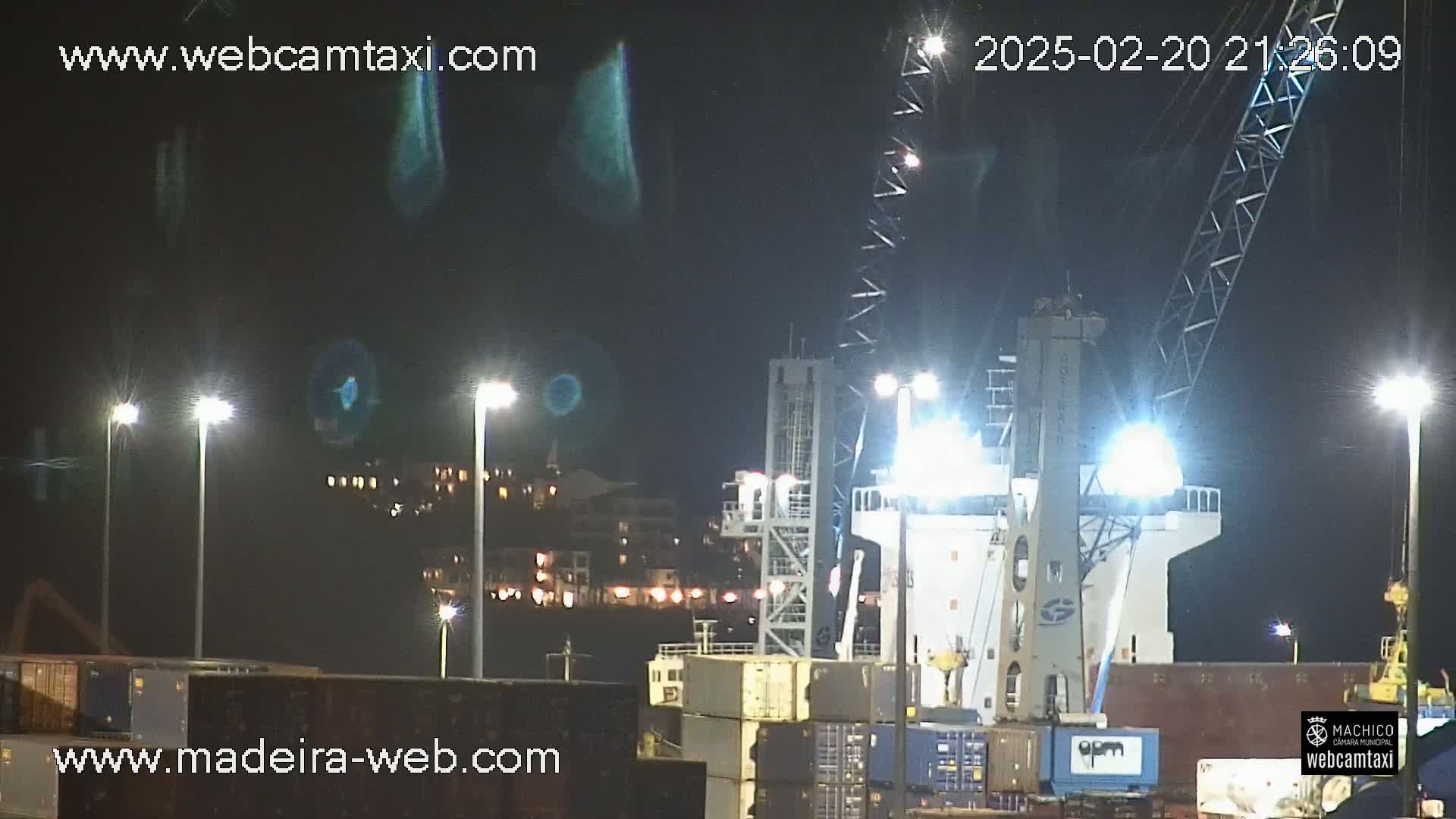At night, a large cargo ship is being loaded at a dock under bright lights.