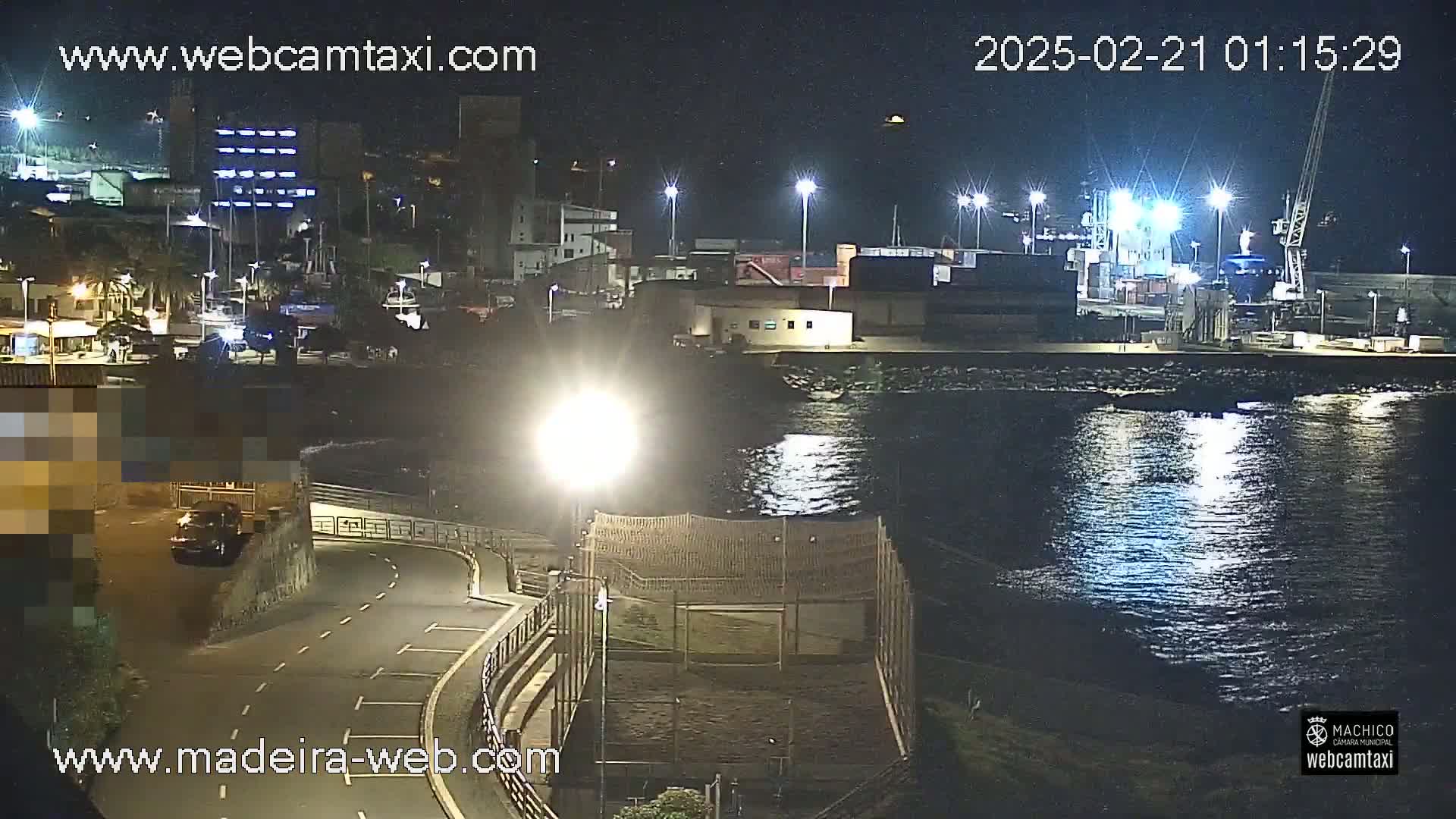 A nighttime view of a harbor with calm water reflecting lights from buildings and docked ships, under a clear sky.