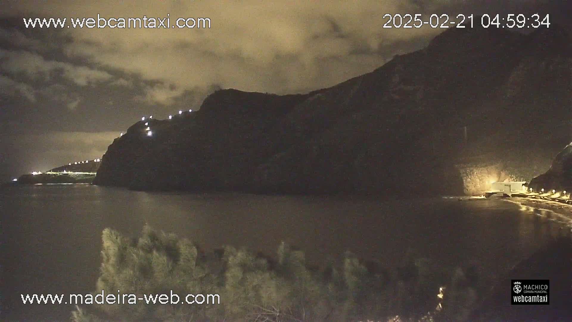 A nighttime view of a dark, rocky coastline with lights visible along the cliff and a calm body of water in the foreground under a cloudy sky.