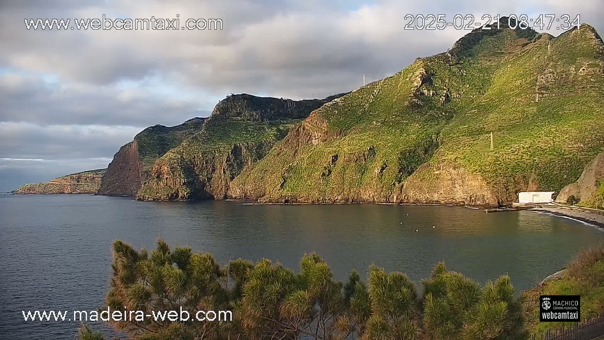 A calm bay is nestled between steep, green cliffs under a mostly cloudy sky.