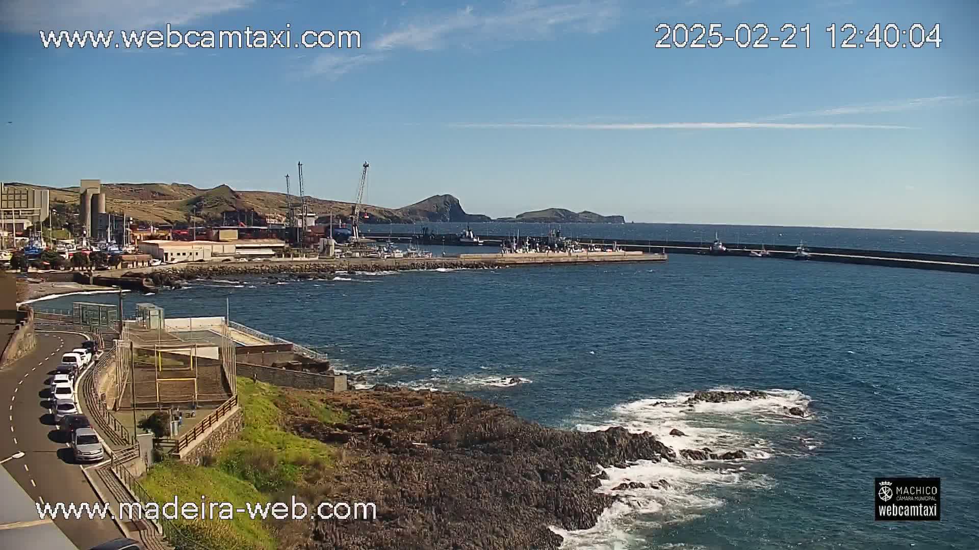 A harbor filled with boats is seen from a high vantage point on a sunny day with a few clouds, overlooking rocky coastlines with some wave action.