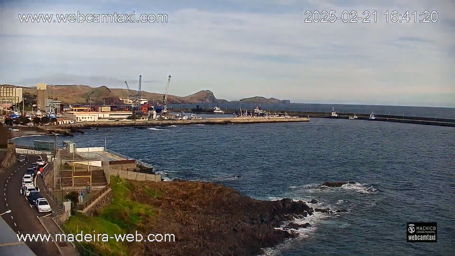 A harbor filled with boats and ships is visible from a rocky shoreline under a partly cloudy sky.