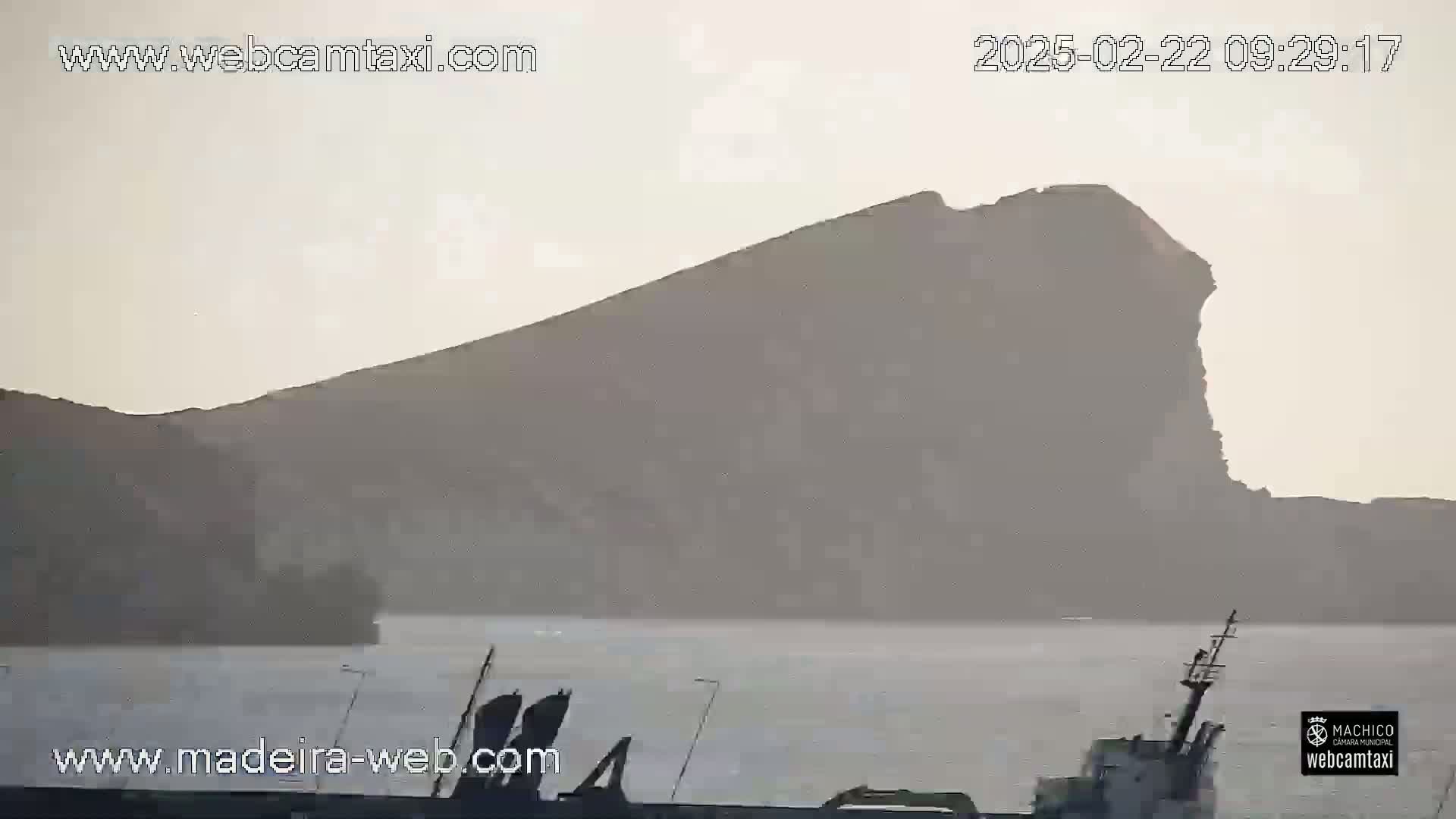 A hazy view shows a large, dark-colored mountain across a body of calm water, with a small boat partially visible in the foreground.