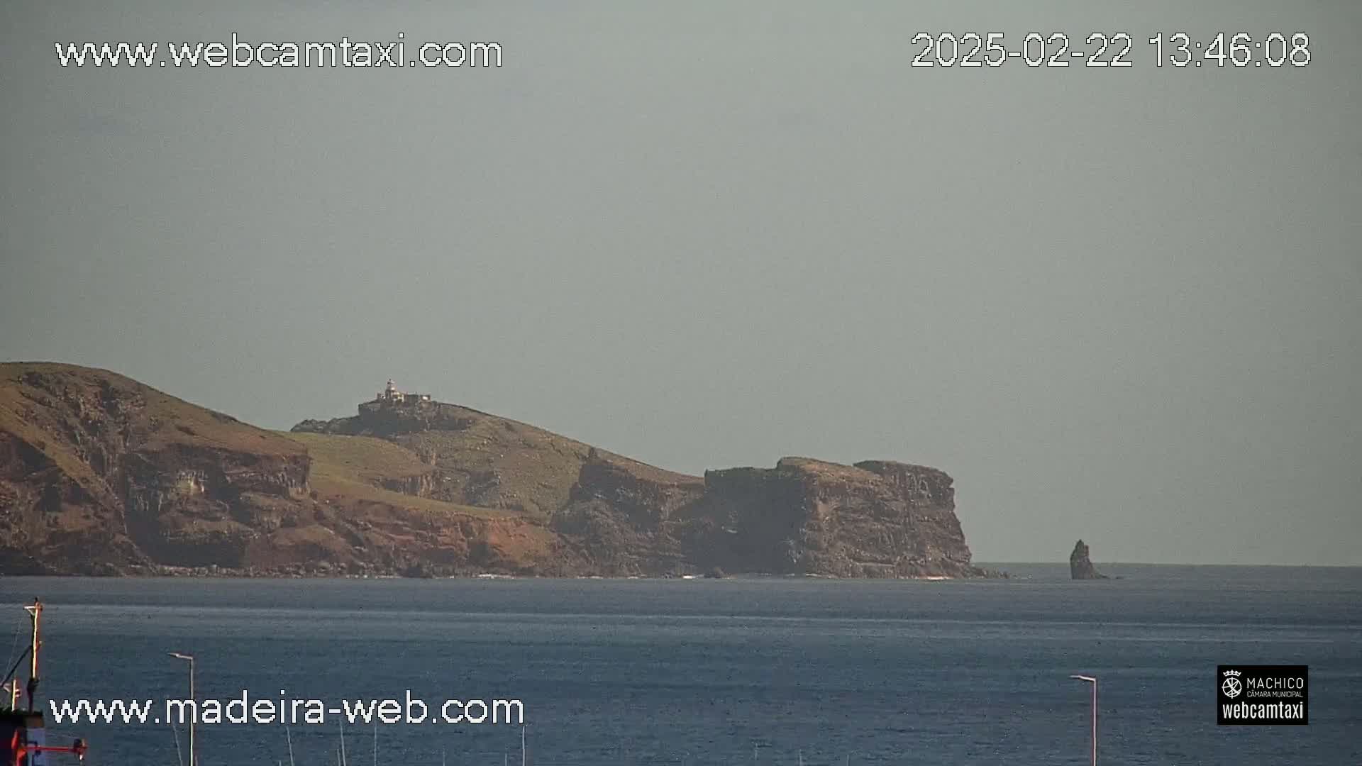 A distant view of a rocky, grassy coastline with a lighthouse, a smaller rock formation in the water, and a hazy sky over calm ocean waters.