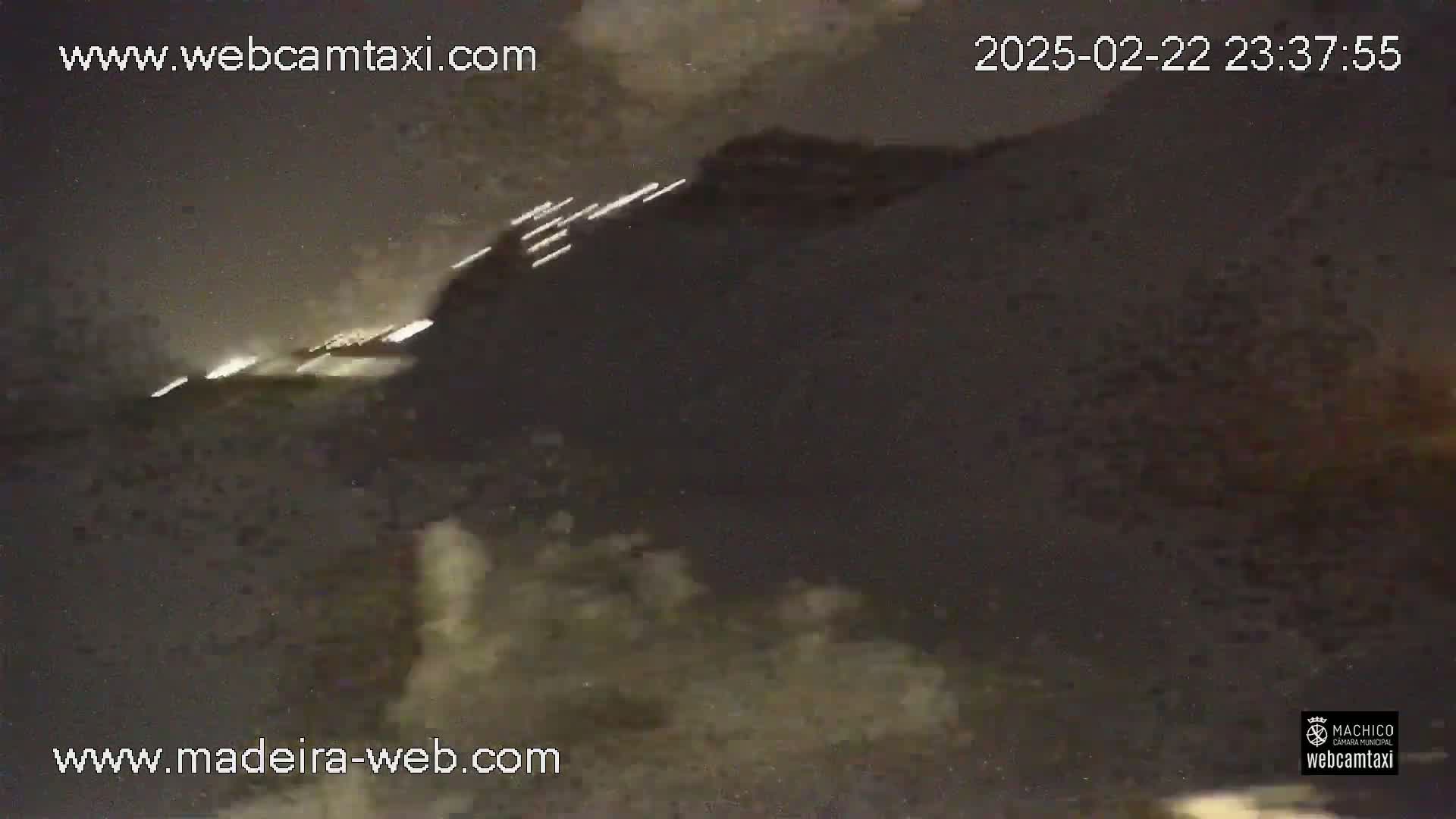 A nighttime, dark-colored, blurry image shows a body of water with some lights visible in the distance and foamy waves near the bottom.