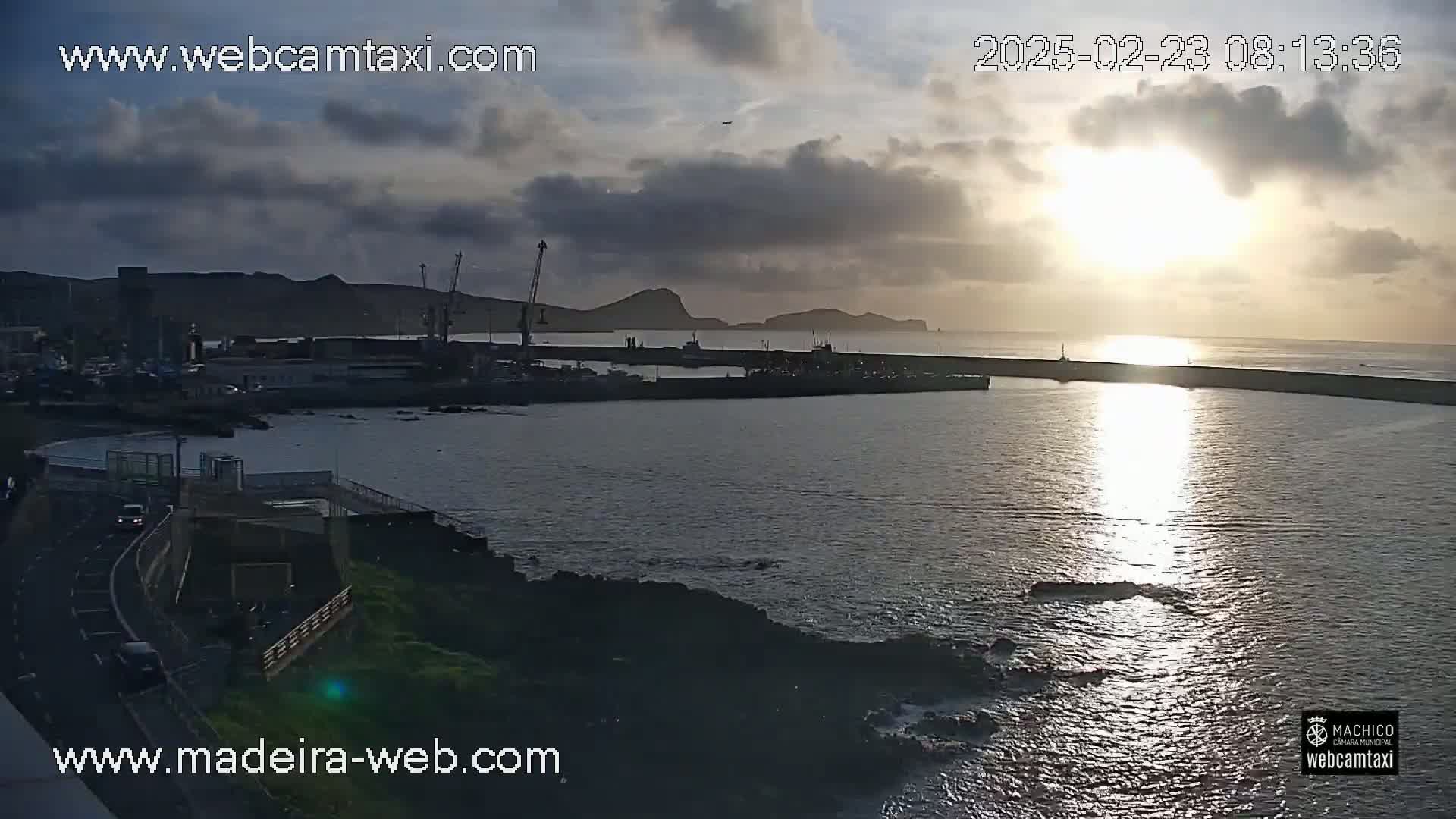A harbor with a breakwater is seen at sunset under a partly cloudy sky.