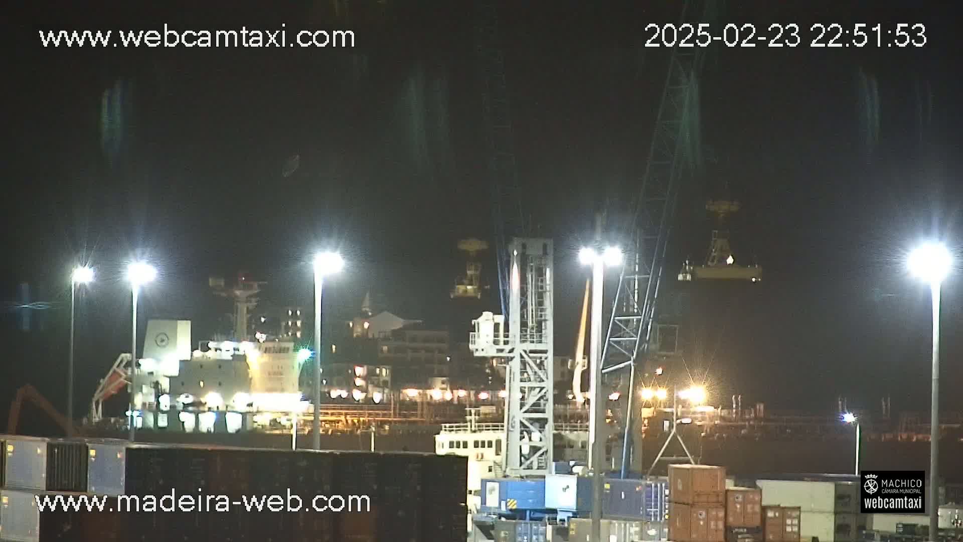 A nighttime view of a cargo ship docked at a port, illuminated by bright lights, under clear skies.