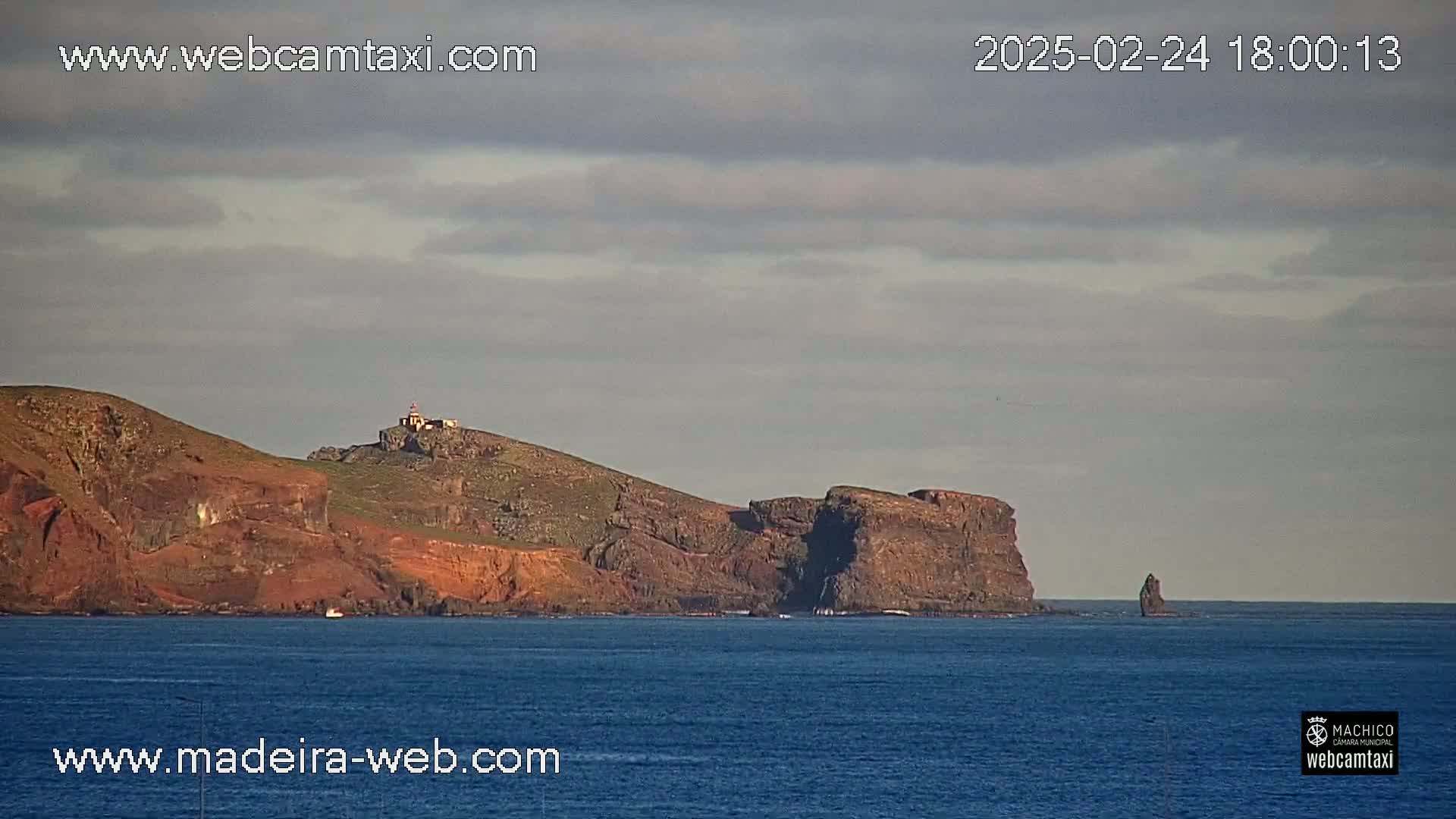 A rocky, reddish-brown coastline with a lighthouse is visible under a mostly cloudy sky over a dark blue ocean.