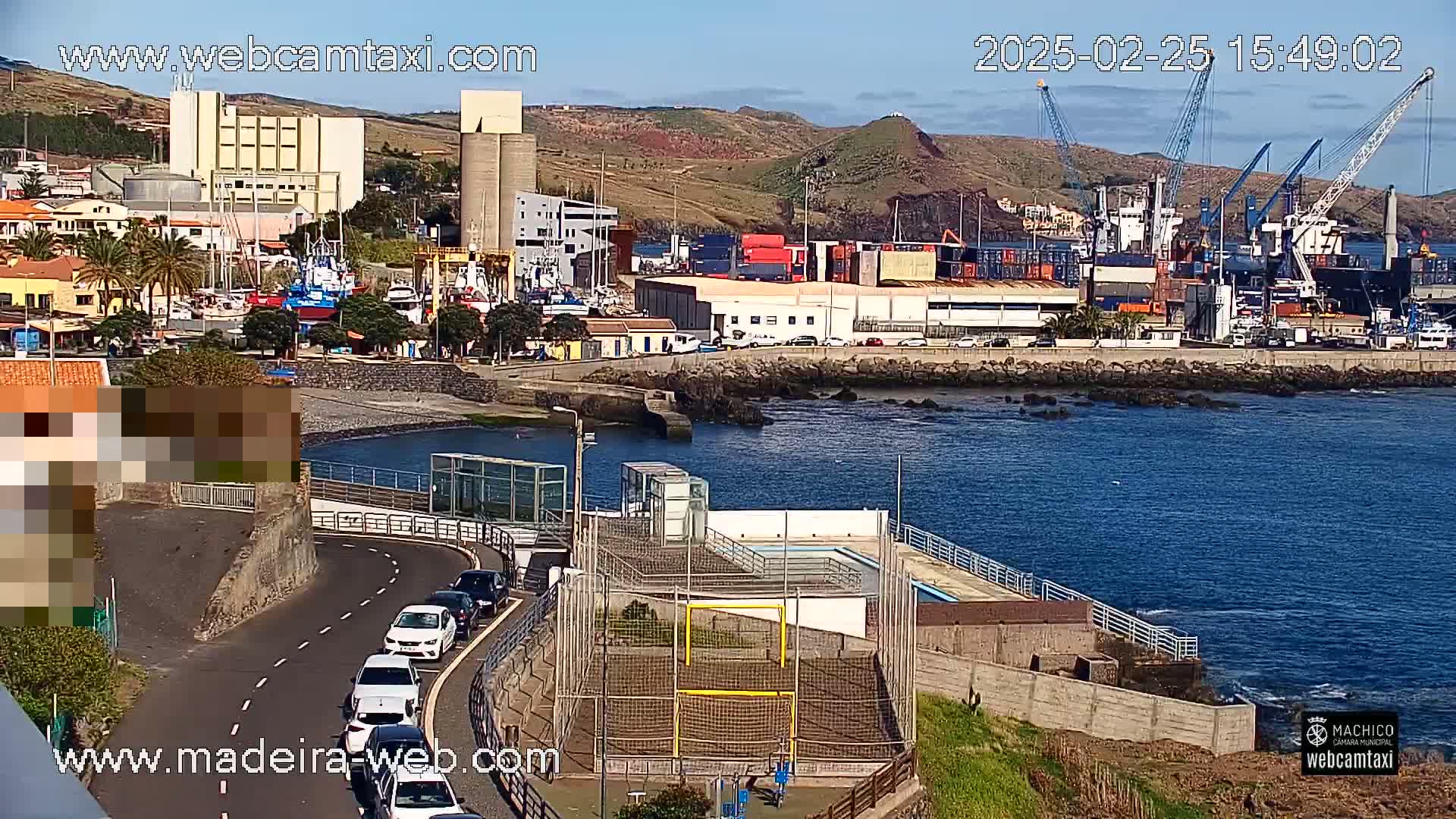 A coastal town's harbor, with a small futsal court near a road, overlooks calm ocean water under a sunny sky.