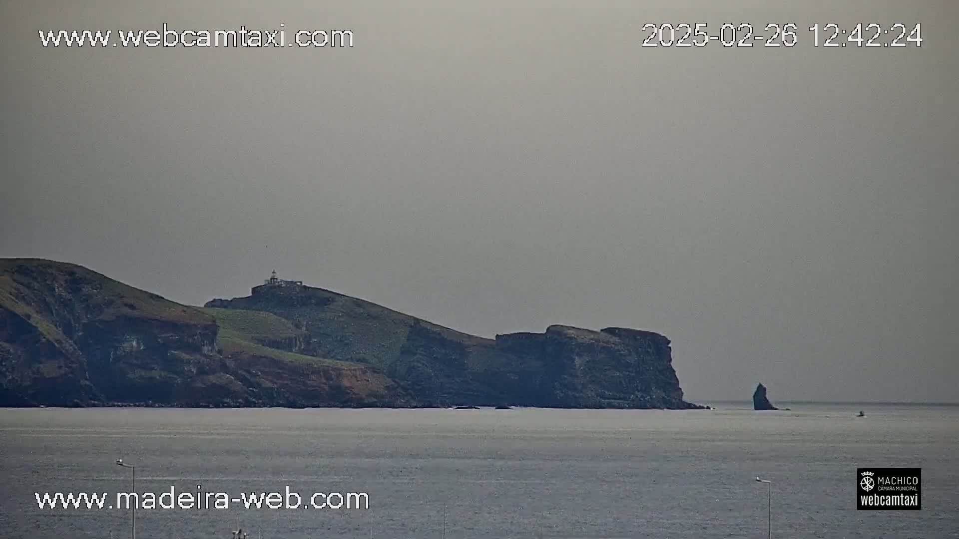A hazy, overcast day reveals a rocky coastal scene with a lighthouse atop a hill, and a solitary rock formation in the calm ocean.
