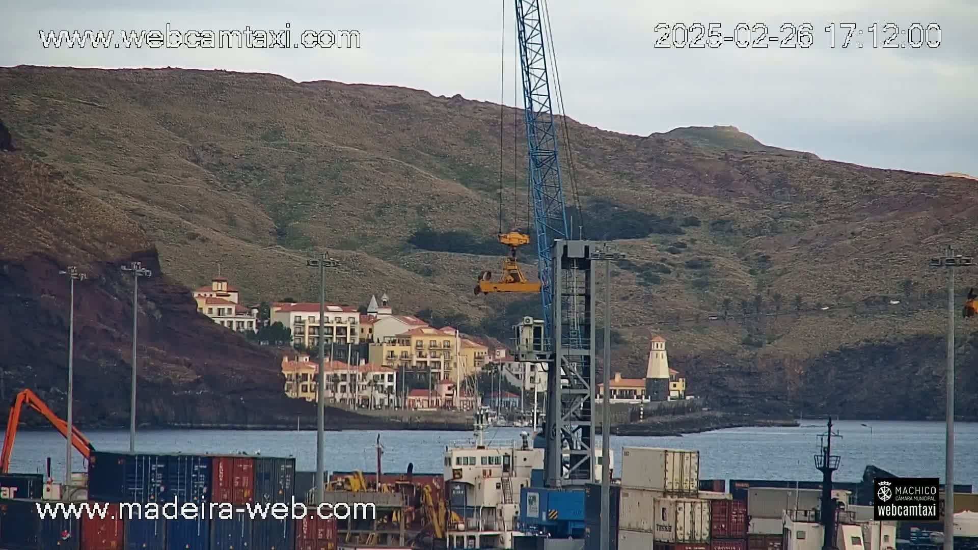 A large blue crane overlooks a harbor filled with cargo containers, with a town nestled against a brown, hilly coastline under an overcast sky.