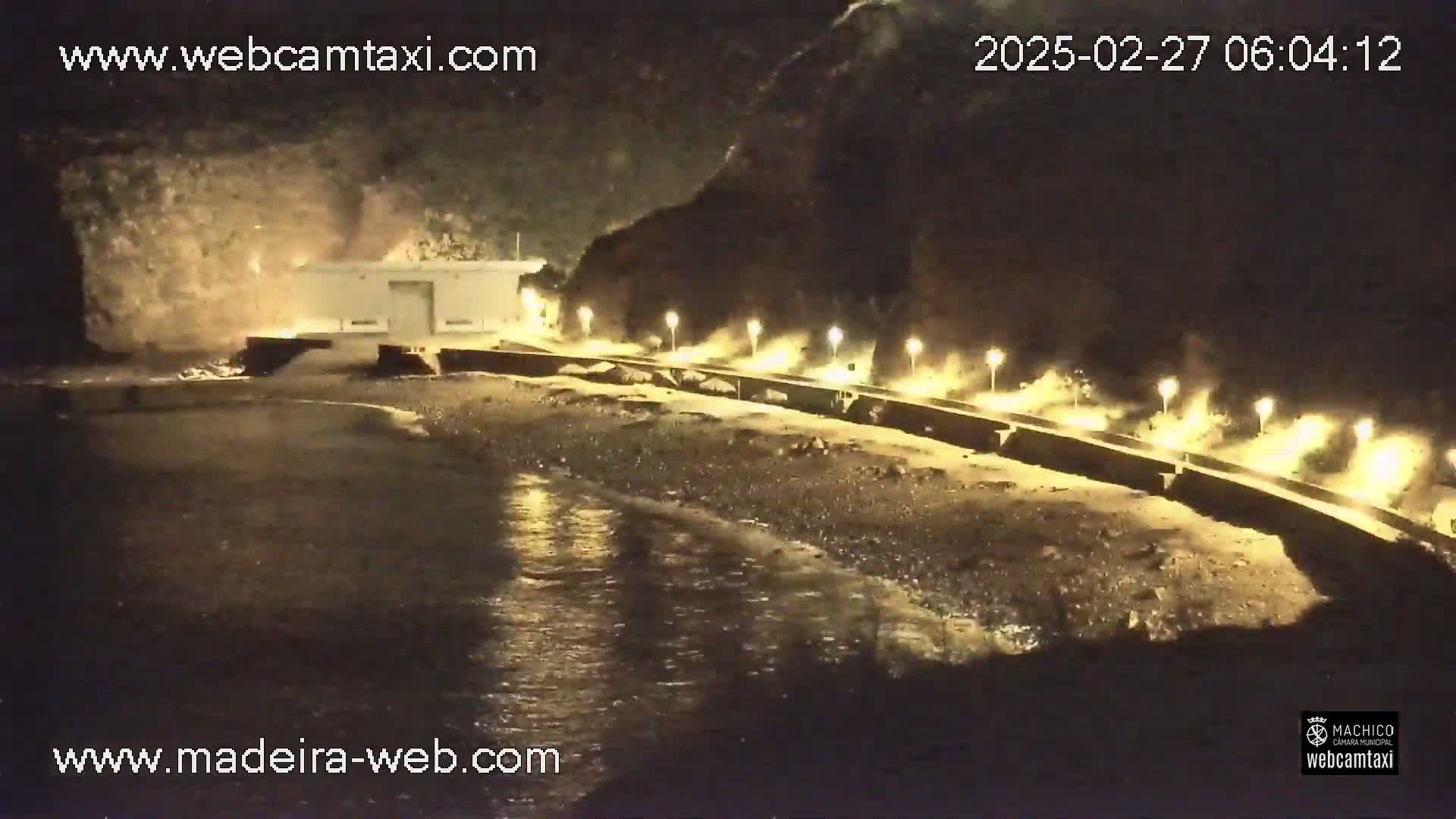 A nighttime coastal scene shows a dark rocky cliff overlooking a beach with a low building and a lighted walkway, and gentle waves lapping the shore.