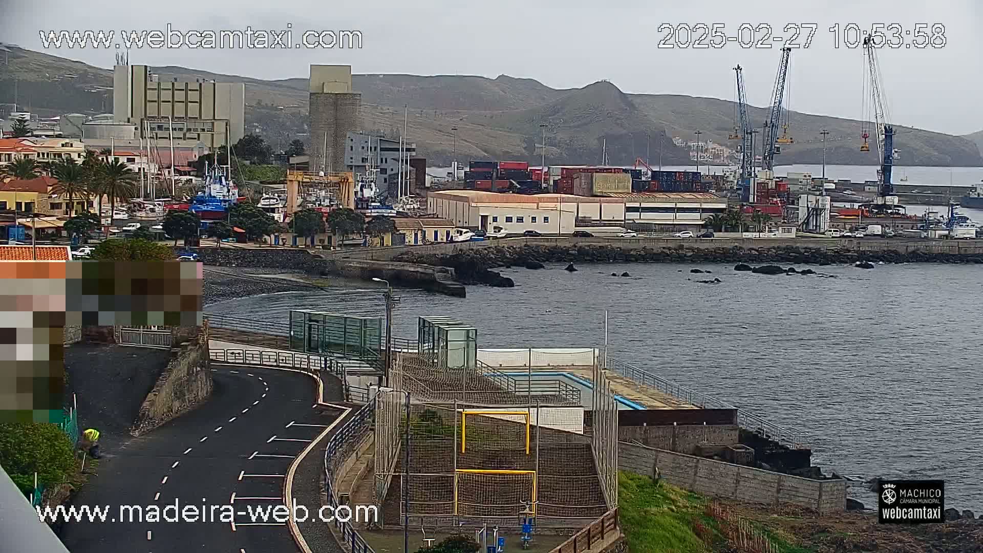 A harbor scene on an overcast day shows a town, a port with cargo containers and cranes, a small beach, and a fenced-in outdoor sports area.