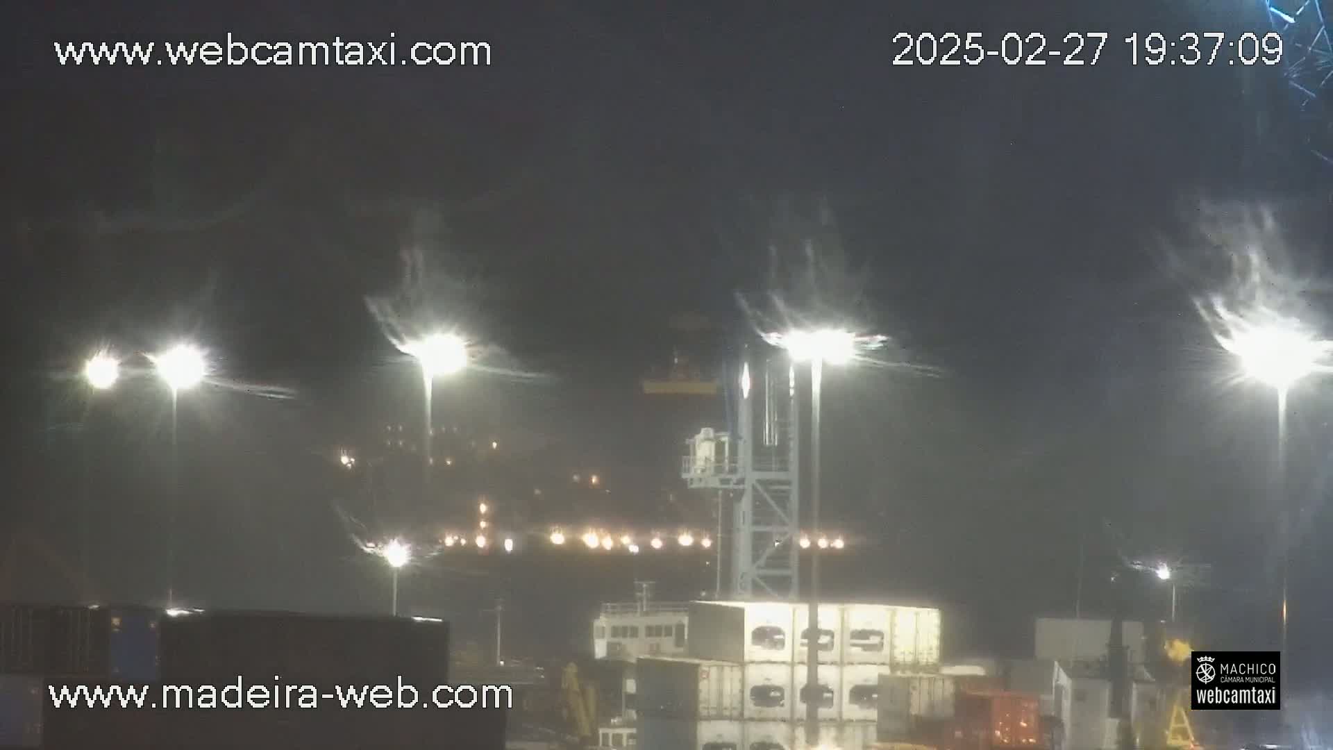 A nighttime view of a port with several tall lights illuminating a foggy or snowy scene, showing containers and a crane.