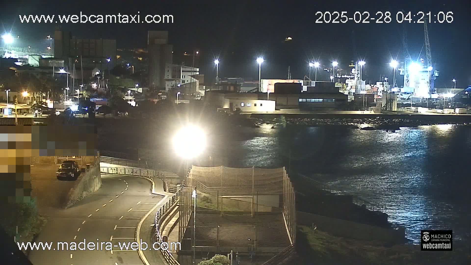 A nighttime view of a harbor with docked ships and boats, illuminated by streetlights, overlooking a calm body of water next to a roadway and a fenced-in area.