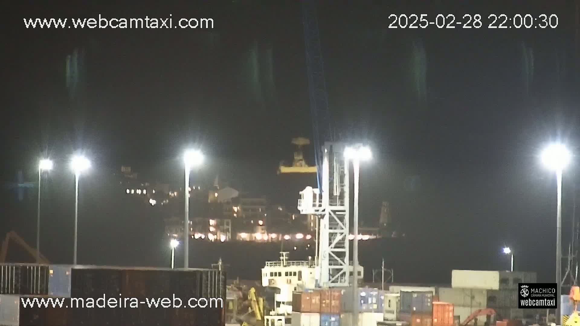 A nighttime view of a port with a large crane and numerous shipping containers, illuminated by bright lights.