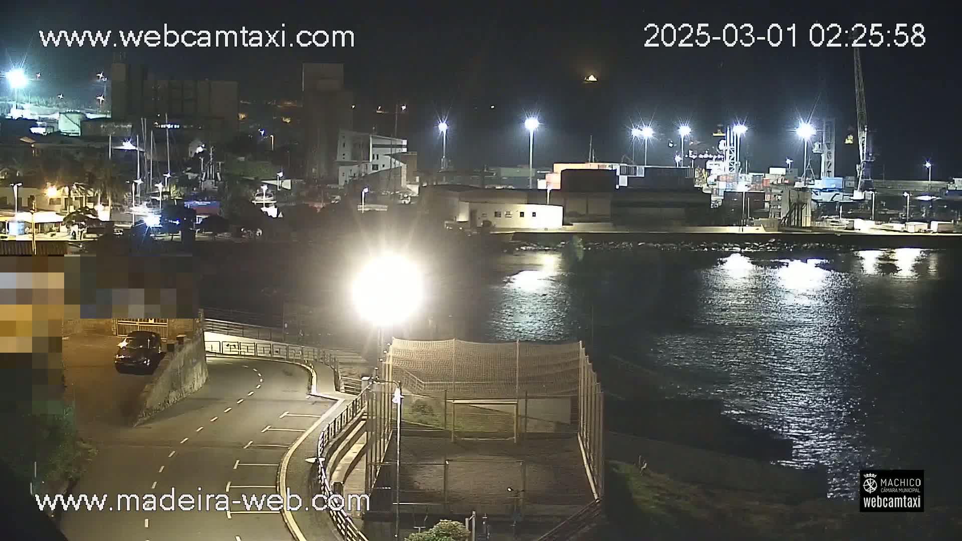A nighttime view of a harbor with a calm body of water, illuminated by numerous lights, and a vehicle parked near a roadway.