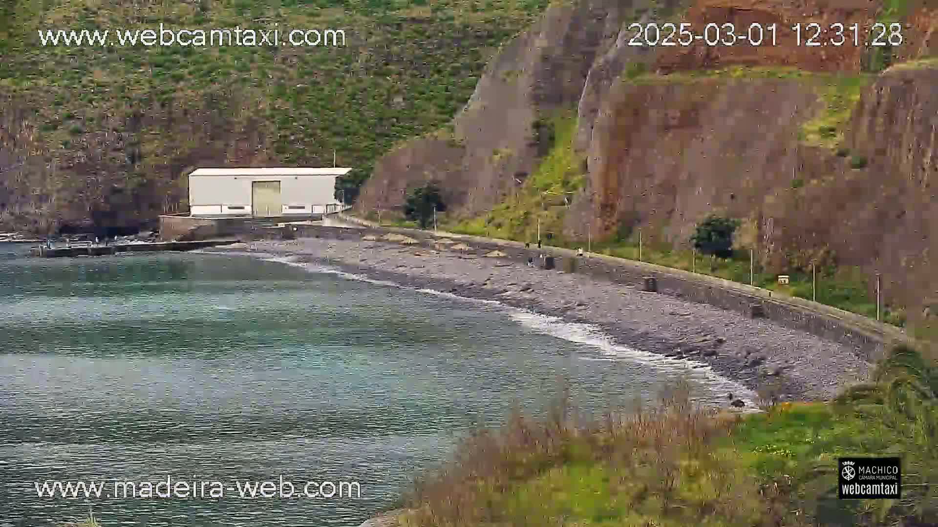 A rocky, dark-colored beach curves along a calm body of water next to a cliffside, with a small white building near the water's edge.