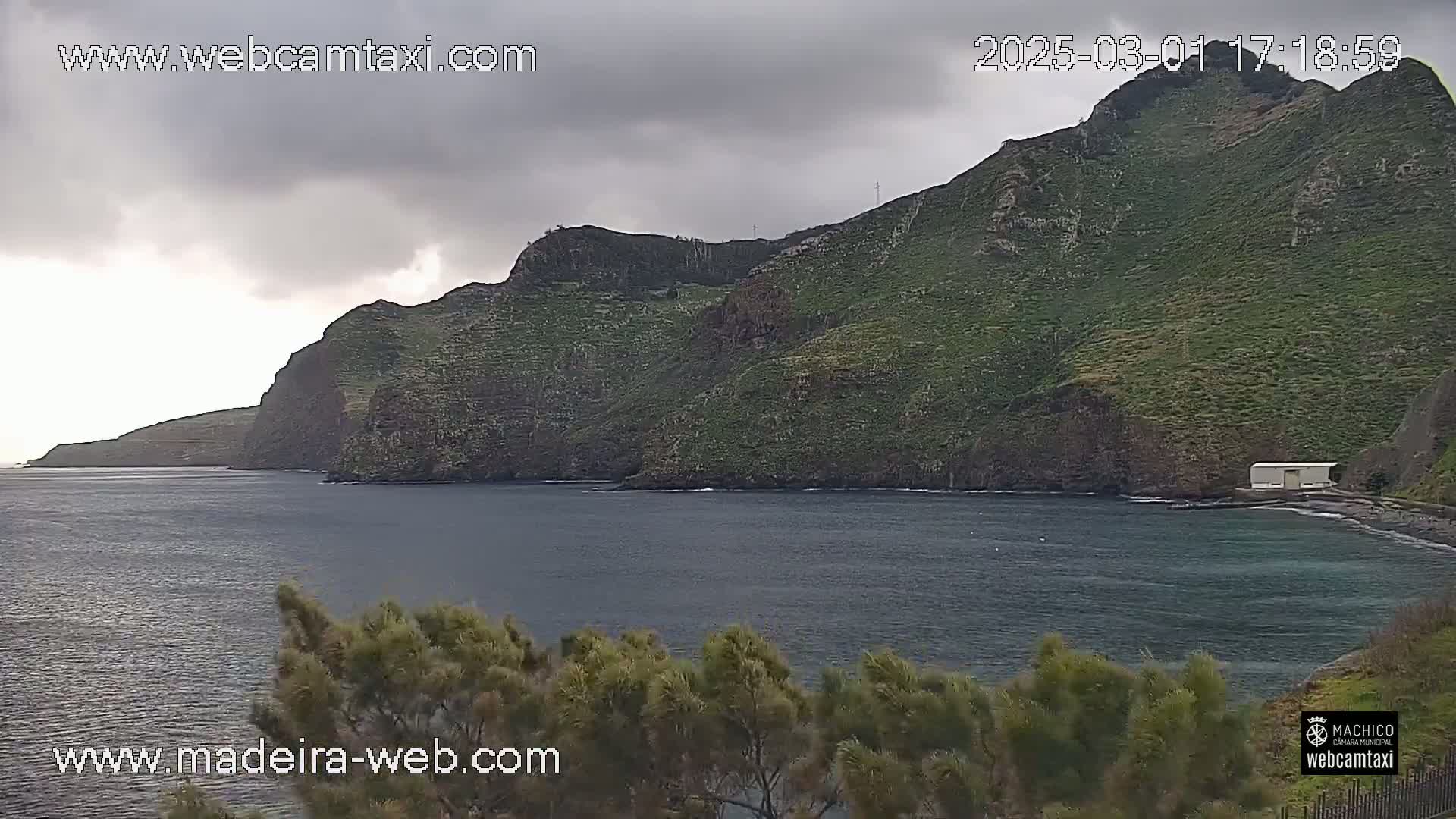 A dark, overcast sky hangs over a rocky, green coastline and a calm, dark-blue ocean.