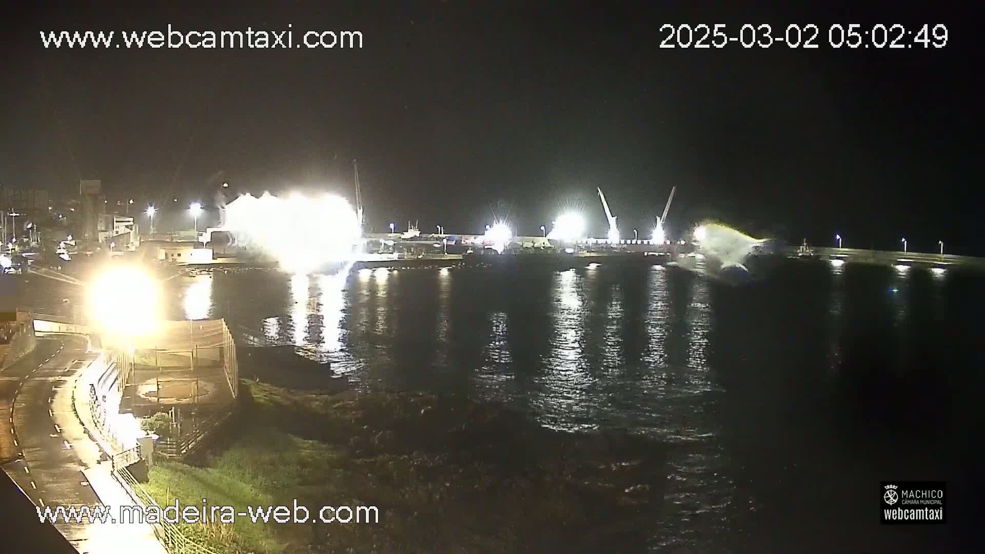 A nighttime harbor scene shows brightly lit industrial activity near the water, with some splashing or spraying visible in the dark water.