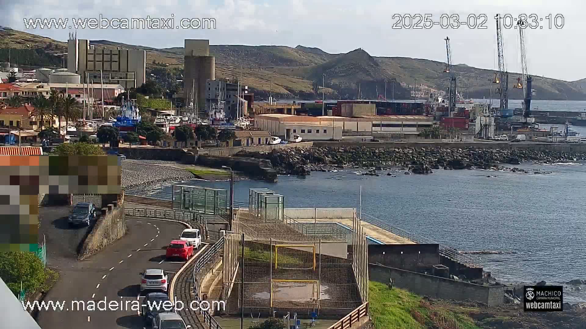 A seaside town with a harbor, industrial buildings, and a small soccer field is visible on a partly cloudy day.