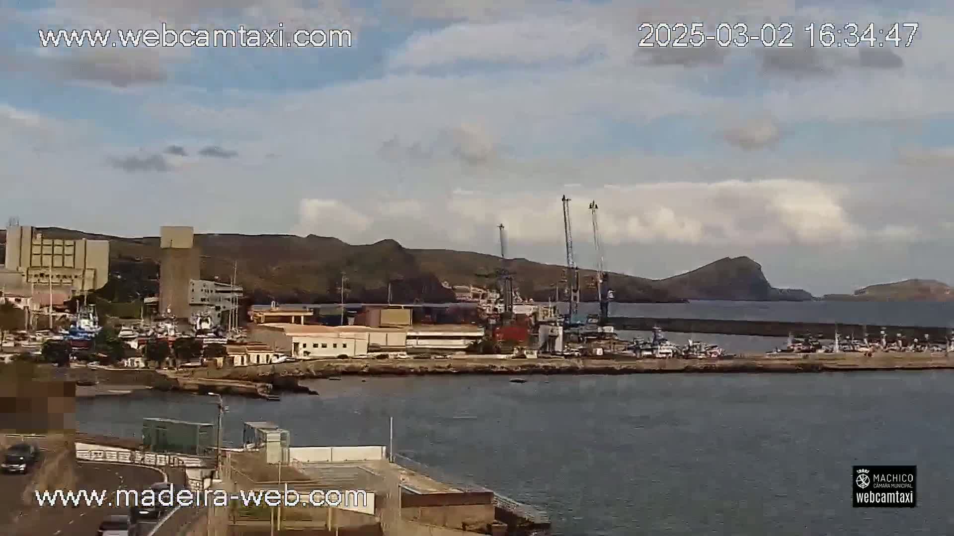A partly cloudy day reveals a harbor filled with boats, industrial buildings, and cranes against a backdrop of hills and calm water.