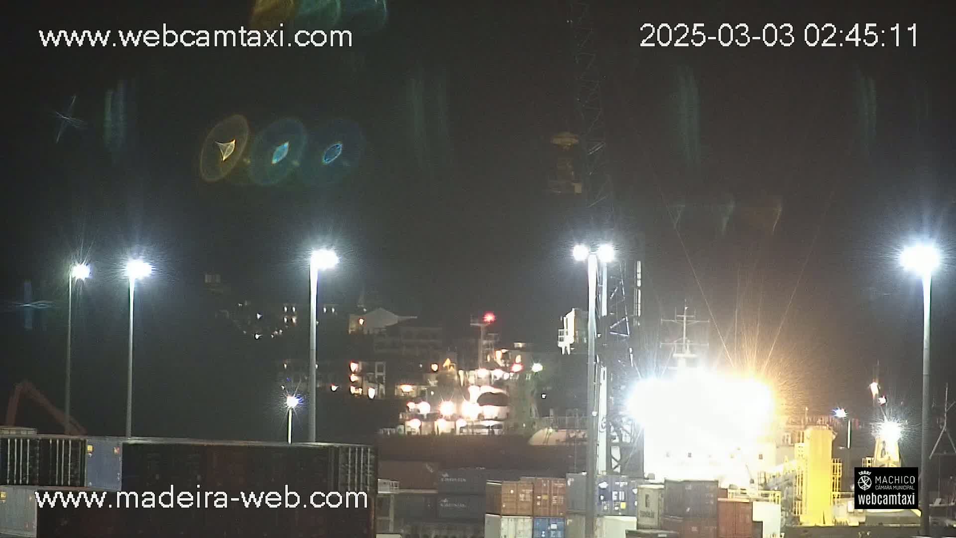 A brightly lit nighttime harbor scene shows a large cargo ship at a dock, with several tall lights illuminating the area.