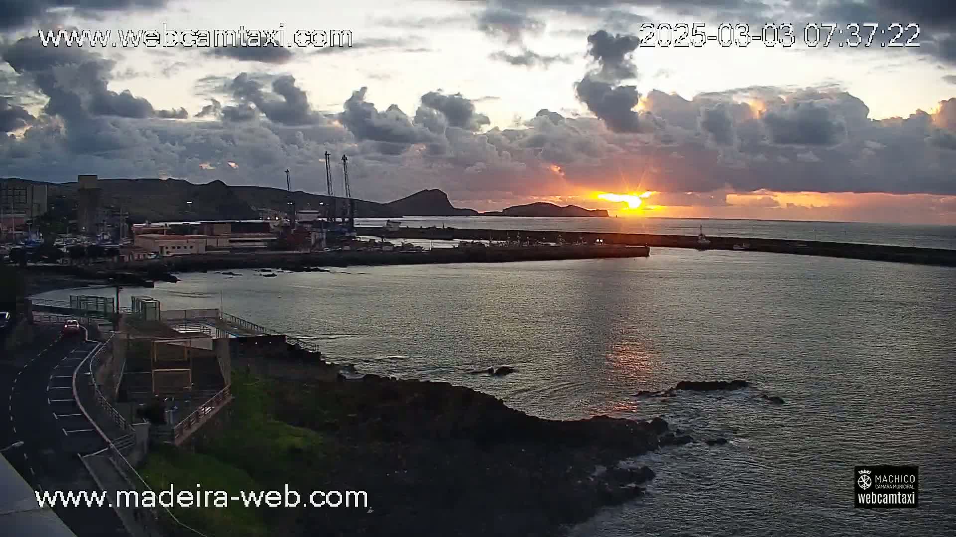A harbor with a breakwater is shown at sunset under a partly cloudy sky.