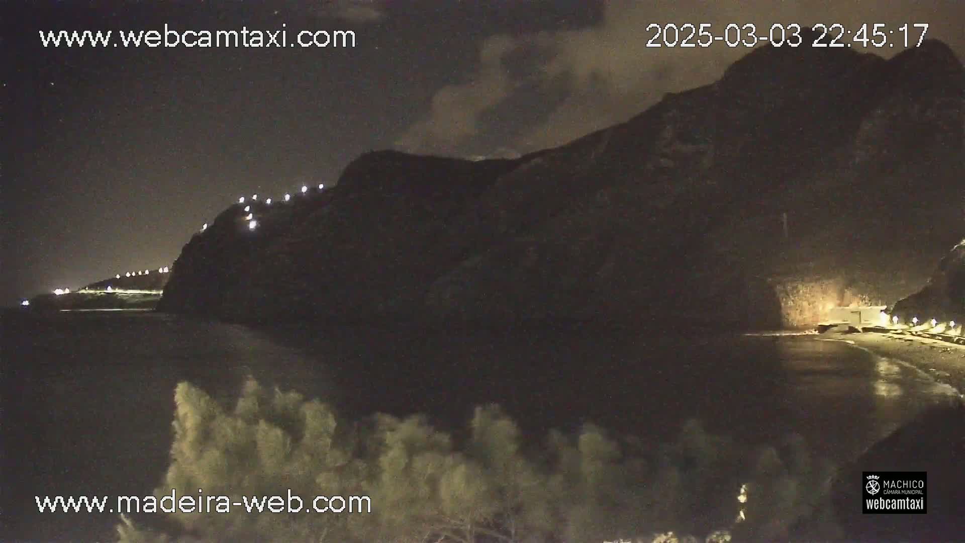 A nighttime coastal scene shows a dark cliff with lights along a road at its top, a dark body of water below, and vegetation in the foreground under a partly cloudy sky.