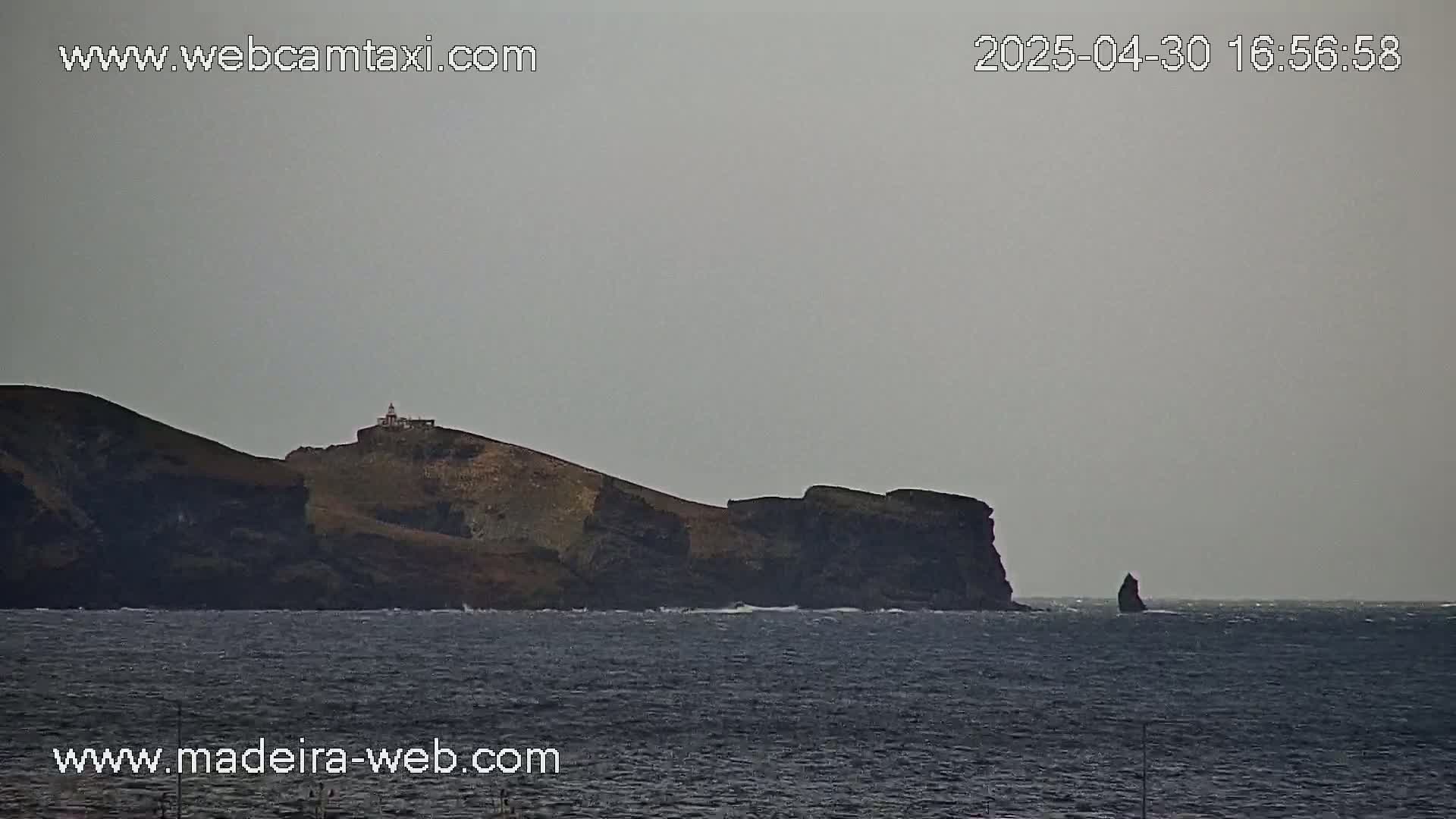 A dark, rocky island with a lighthouse sits on the horizon of a calm, gray ocean under an overcast sky.