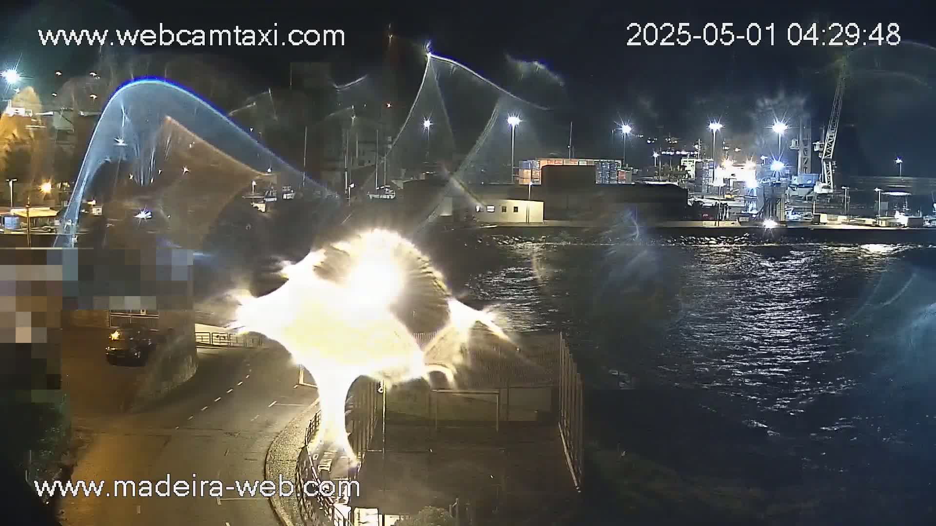 A nighttime view of a harbor with choppy water, illuminated by streetlights, features a bright, indistinct object near the shore.