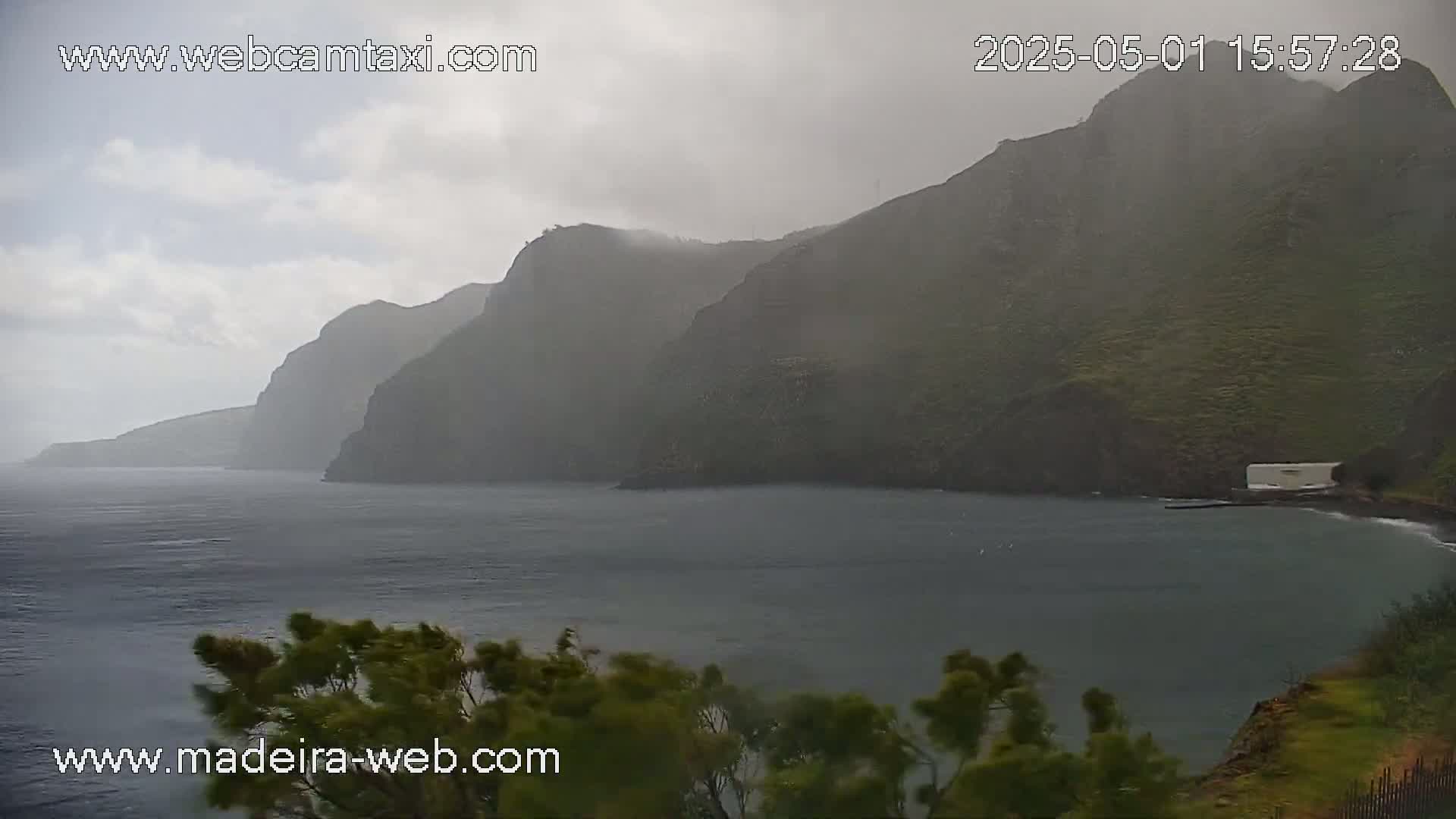 Rain falls on a dark, rocky coastline with a calm body of water in the foreground.