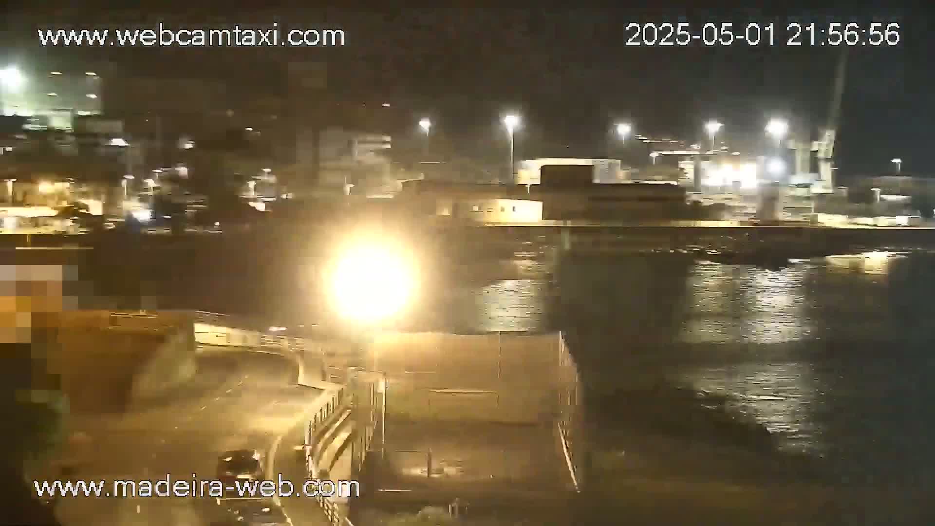 A nighttime view of a harbor with calm water, illuminated by lights from buildings and streetlights on a roadway, shows some vehicles on the road.