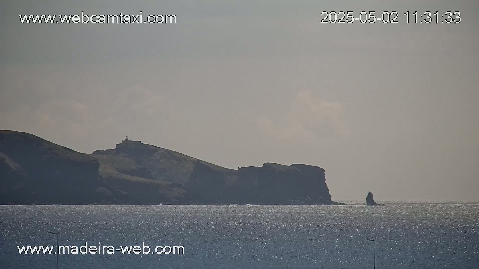 A hazy, sunny day reveals a dark, rocky coastline with a lighthouse atop a hill, a smaller rock formation in the water, and a calm sea.