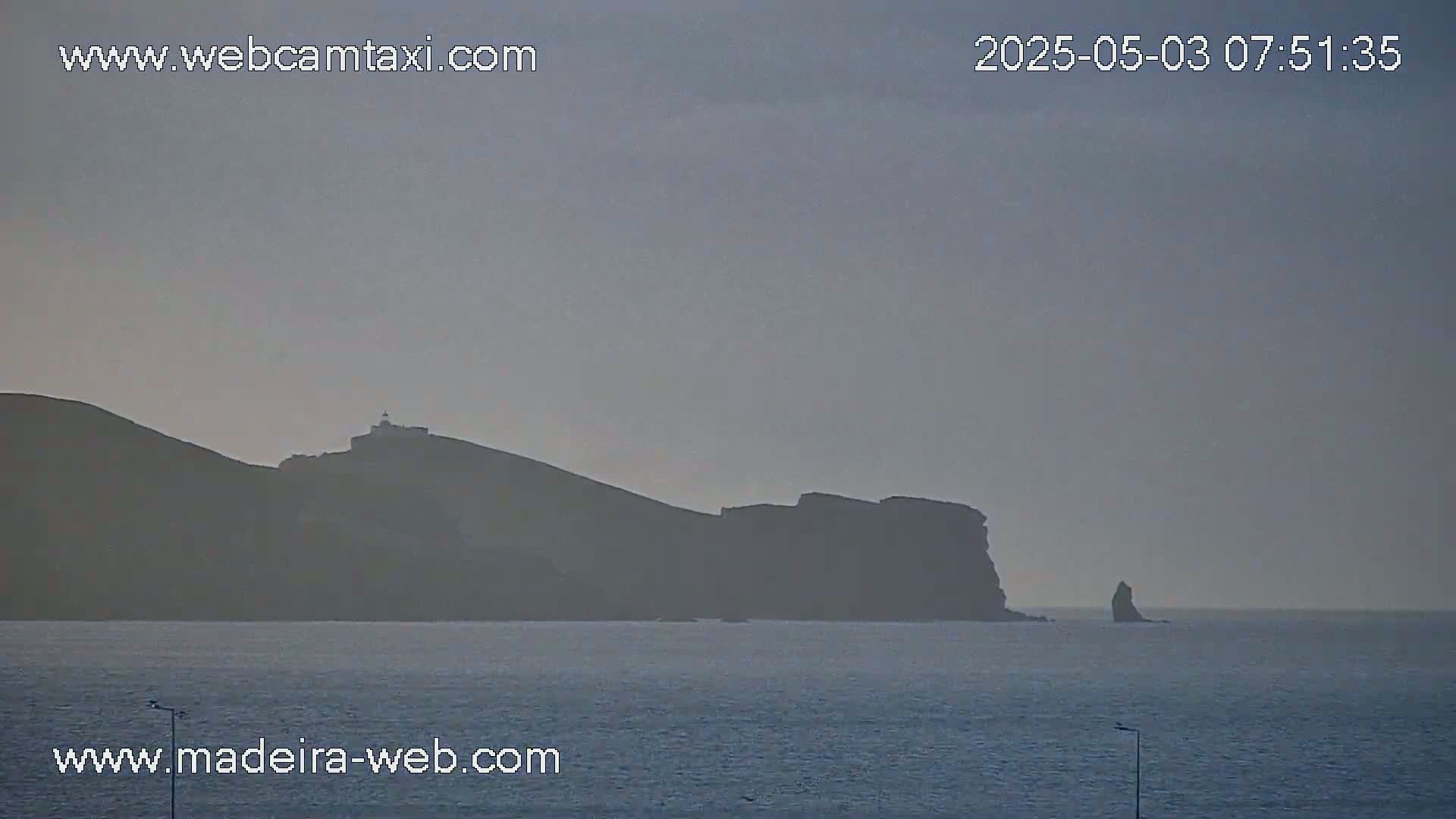 A hazy view shows a dark, rocky coastline with a lighthouse, extending into a calm, gray sea.