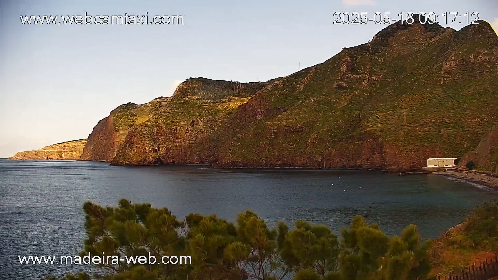 A rocky, green-brown cliffside overlooks a calm, dark-blue bay under a clear, light-blue sky.