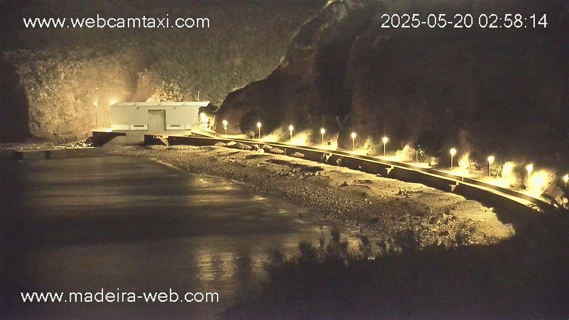 A nighttime view of a beach with a building, a paved walkway lined with lights, and a rocky shoreline under a clear sky.