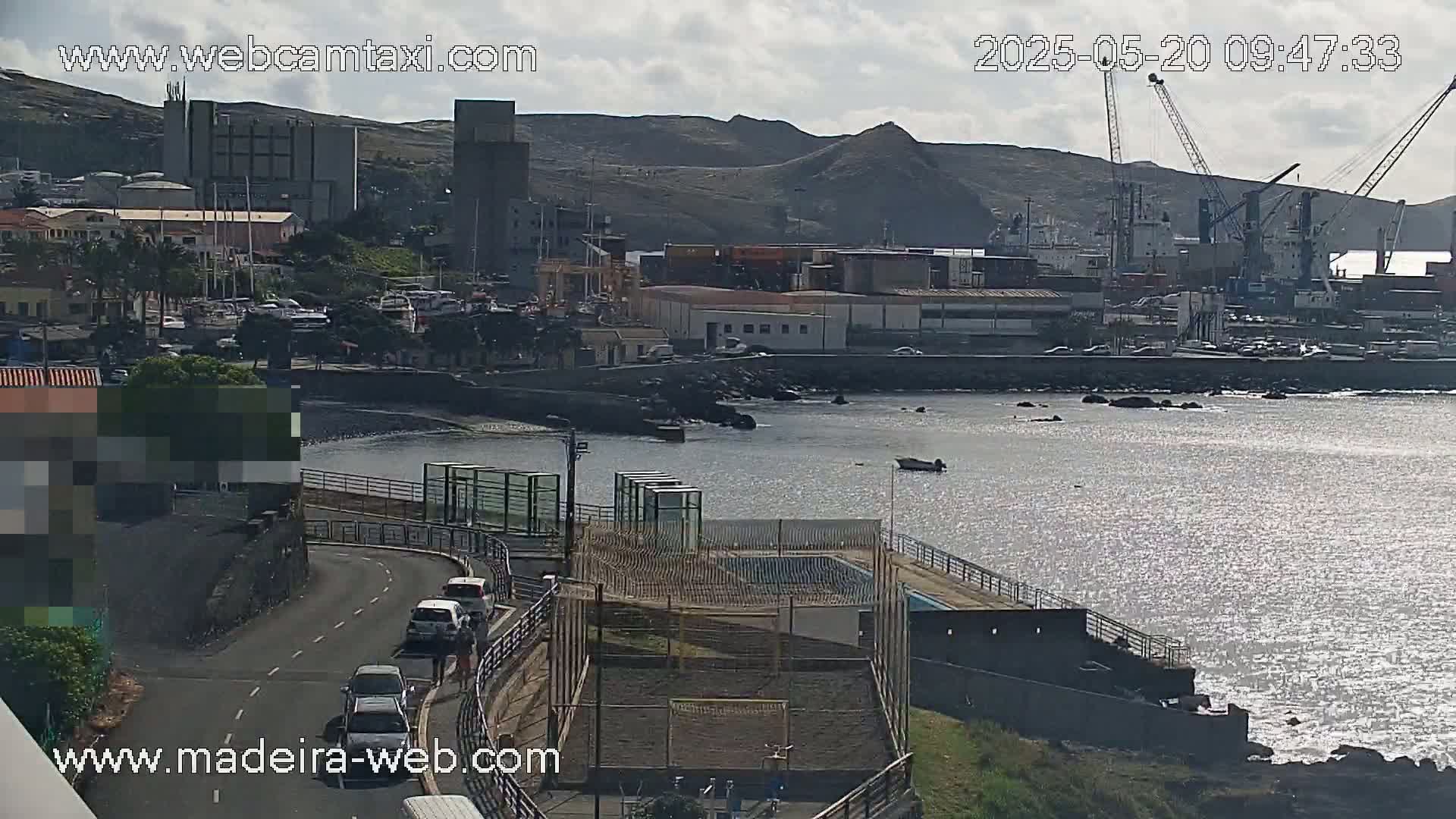 A coastal town with a harbor, industrial buildings, and several vehicles on a road overlooking a calm, sunlit body of water under a partly cloudy sky.