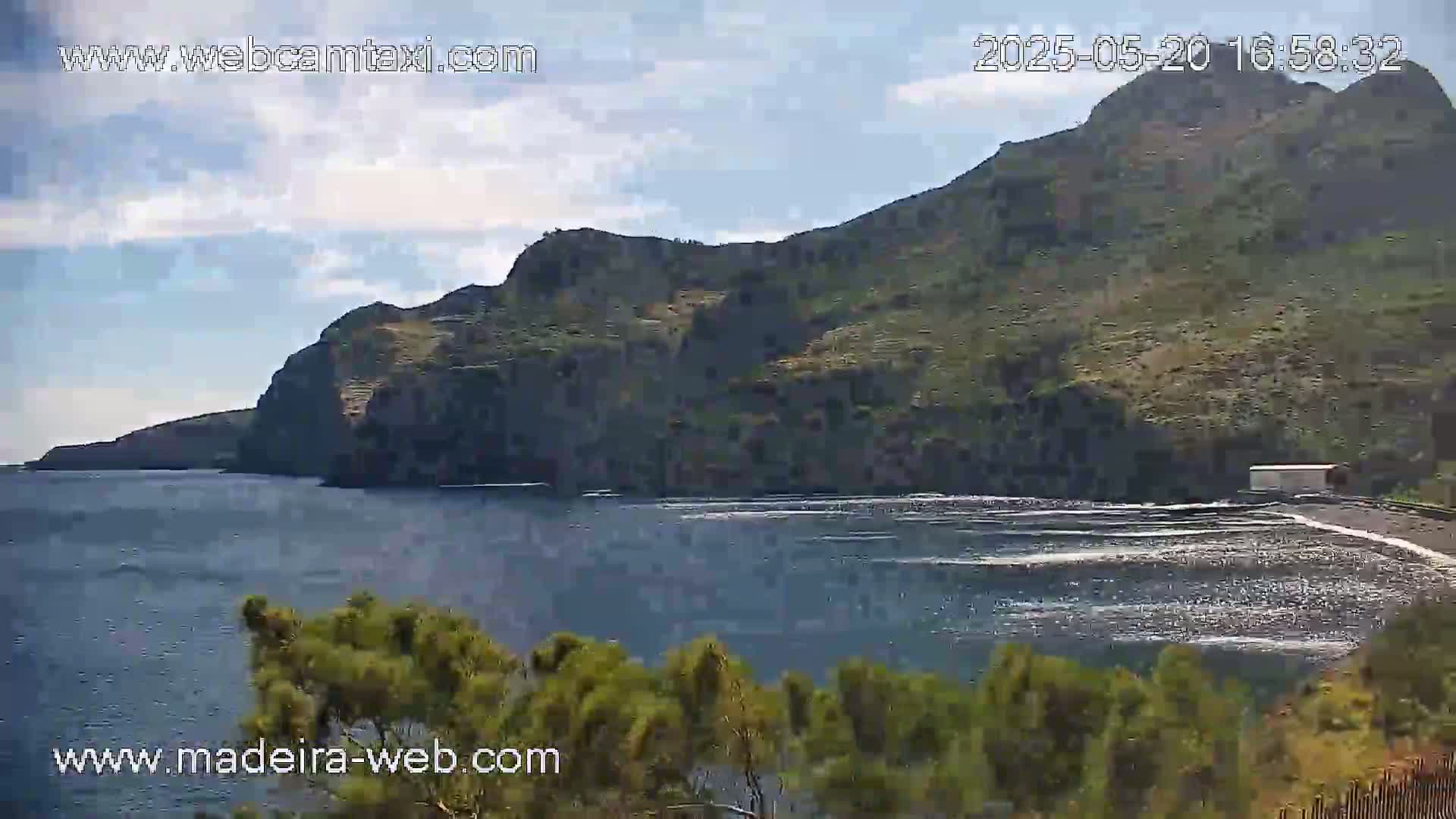 A dark-colored, rocky coastline meets a calm ocean under a partly cloudy sky.